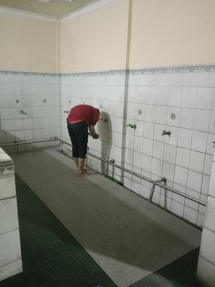 The image shows a man in a red shirt and black pants washing his hands in a tiled washroom. He is standing at a tiled sink with a stainless steel handrail. The room is located in a building in Tugu, Indonesia, at the coordinates -6.976, 110.331. There is a gray carpet on the floor and a green mat on the floor in the lower left corner. There are a few taps on the wall above the sink. The room has white walls and a white tiled floor.