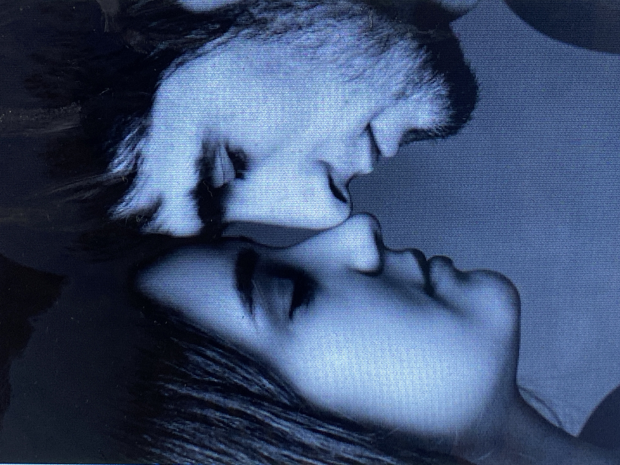 The image is a close-up, blue-toned photograph of two people, likely a young couple, in an intimate pose. Their foreheads are touching, and their noses are nearly touching, suggesting a moment of deep connection or a kiss. The woman is on the left, with her eyes closed and her head slightly tilted. She has dark hair. The man is on the right, with his eyes also closed and a slight smile. He has darker, unkempt hair and some facial hair. The background is a dark, undifferentiated color, emphasizing the subjects. There are no visible details of a specific city or location, including Marghedar, Tajikistan.