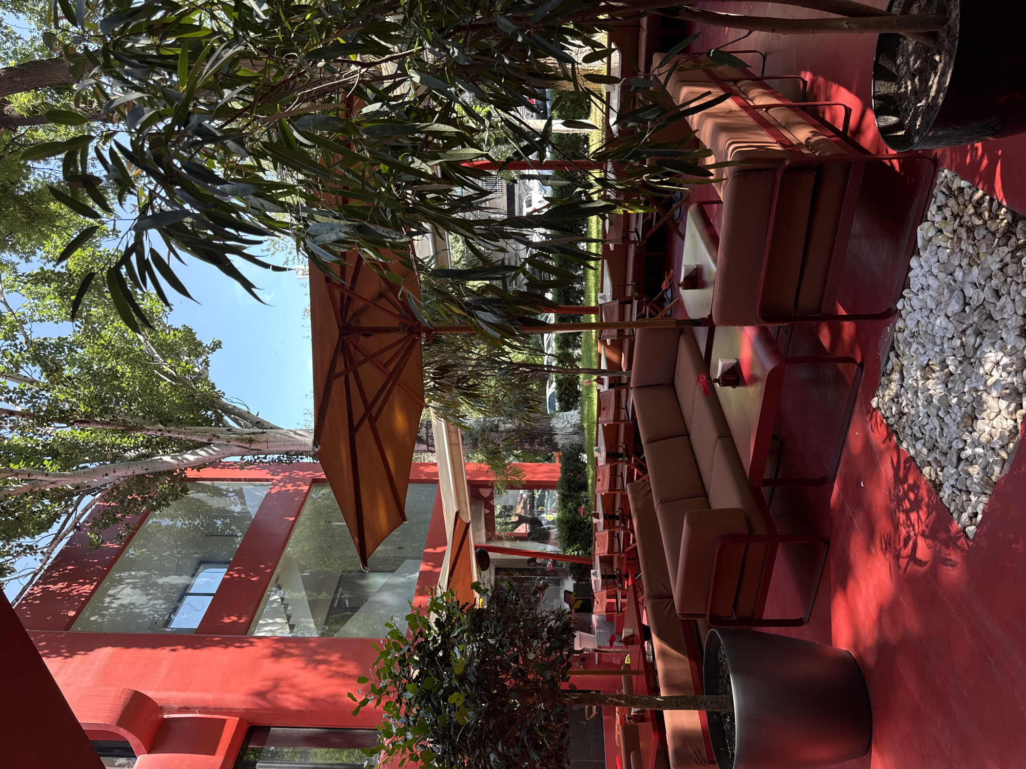 This is an outdoor seating area of a modern restaurant, likely in Yerevan, Armenia, given the architectural style. The entire area is painted a vibrant red, including the floor, tables, chairs, and the exterior of the building. Large, contemporary sofas and armchairs upholstered in a brown fabric are arranged around red coffee tables. Several large umbrellas, also in a reddish-brown hue, provide shade. Potted plants, including a large tree with dark green, elongated leaves and a smaller, bush-like tree, are placed throughout the seating area. The red building behind the seating area features large, floor-to-ceiling windows. Trees with green foliage are visible in the background, and a bright blue sky is present. The scene suggests a well-maintained, stylish outdoor dining space.