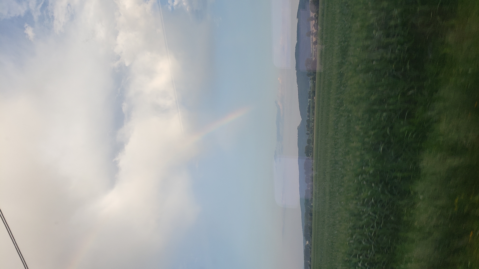 The image depicts a rural landscape under a partly cloudy sky. A faint rainbow is visible in the upper portion of the frame, arcing from the clouds towards the horizon. Below the sky, a wide expanse of green field stretches across the foreground and midground. In the distance, a tree line and rolling hills form the backdrop, with some structures suggesting a small settlement or farmsteads. The overall impression is one of natural scenery, possibly captured during or after a rain shower. The location is identified as Poprad, Slovakia.