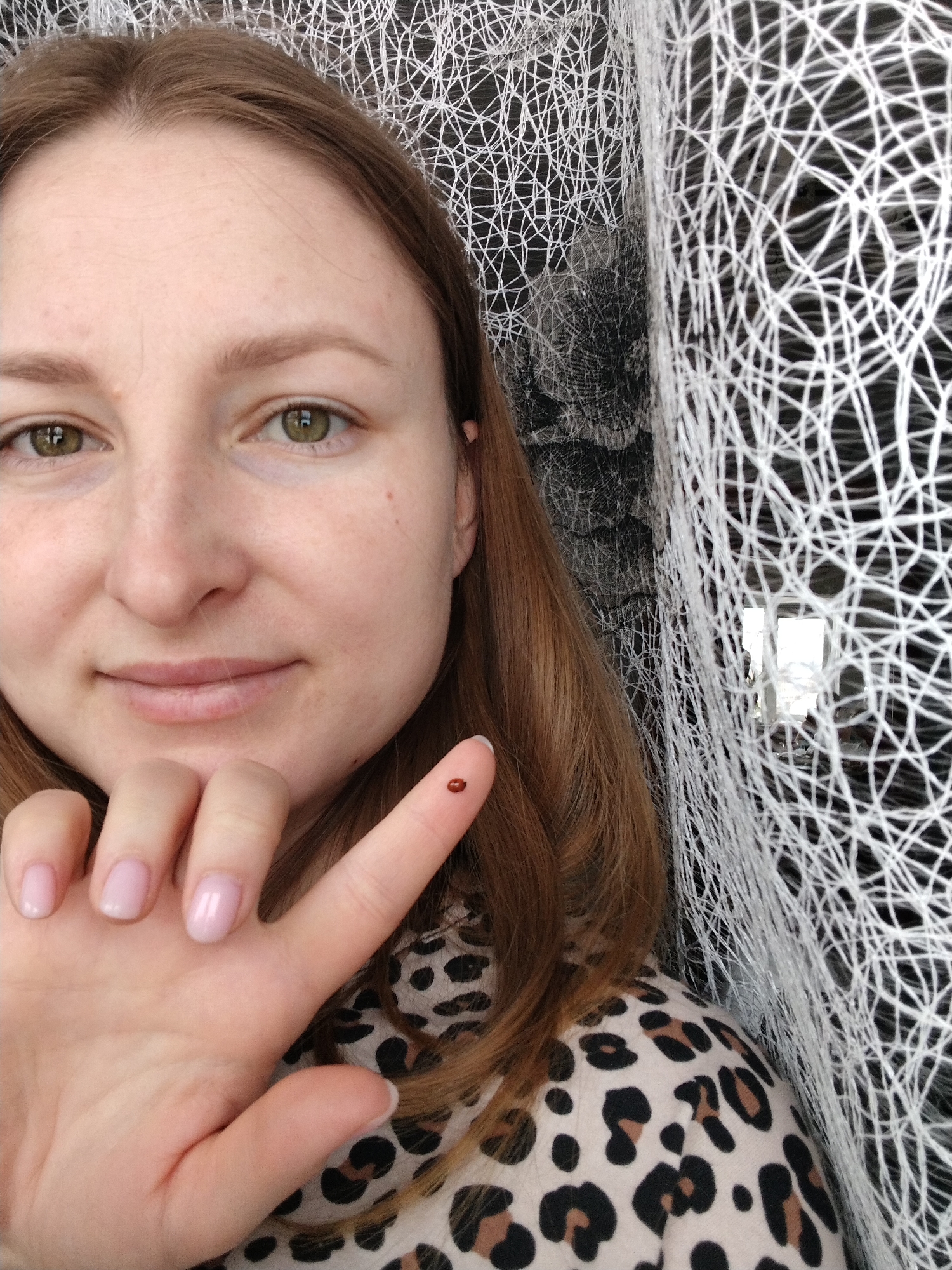 A female individual, appearing Caucasian with hazel eyes and medium-brown hair, is depicted in an indoor environment in Nizhnyaya Gar', Russia. She faces the viewer with a slight smile. Her left hand is prominently featured, with a small, reddish-brown, oval-shaped insect, consistent with a ladybug, resting on the tip of her index finger. Her fingernails are short and painted a pale pink. She wears a cream-colored garment with a black and brown leopard print. The background consists of a white, irregularly woven mesh fabric, behind which a dark surface is visible. A rectangular reflection, indicative of an interior window or light fixture, is partially obscured by the mesh on the right side of the frame.