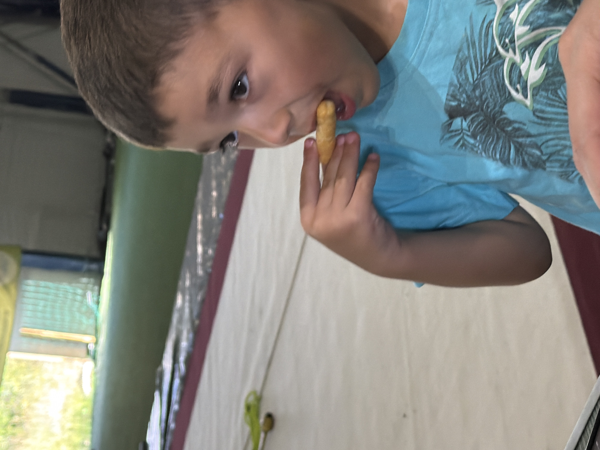 In Sofia, Bulgaria, a young boy with brown hair is in an indoor sports facility. He is wearing a blue t-shirt with a palm tree and dolphin design. The boy is holding a snack to his mouth and appears to be eating it. The background shows a green sports turf area and some fencing. The flooring is a light-colored surface with red trim.