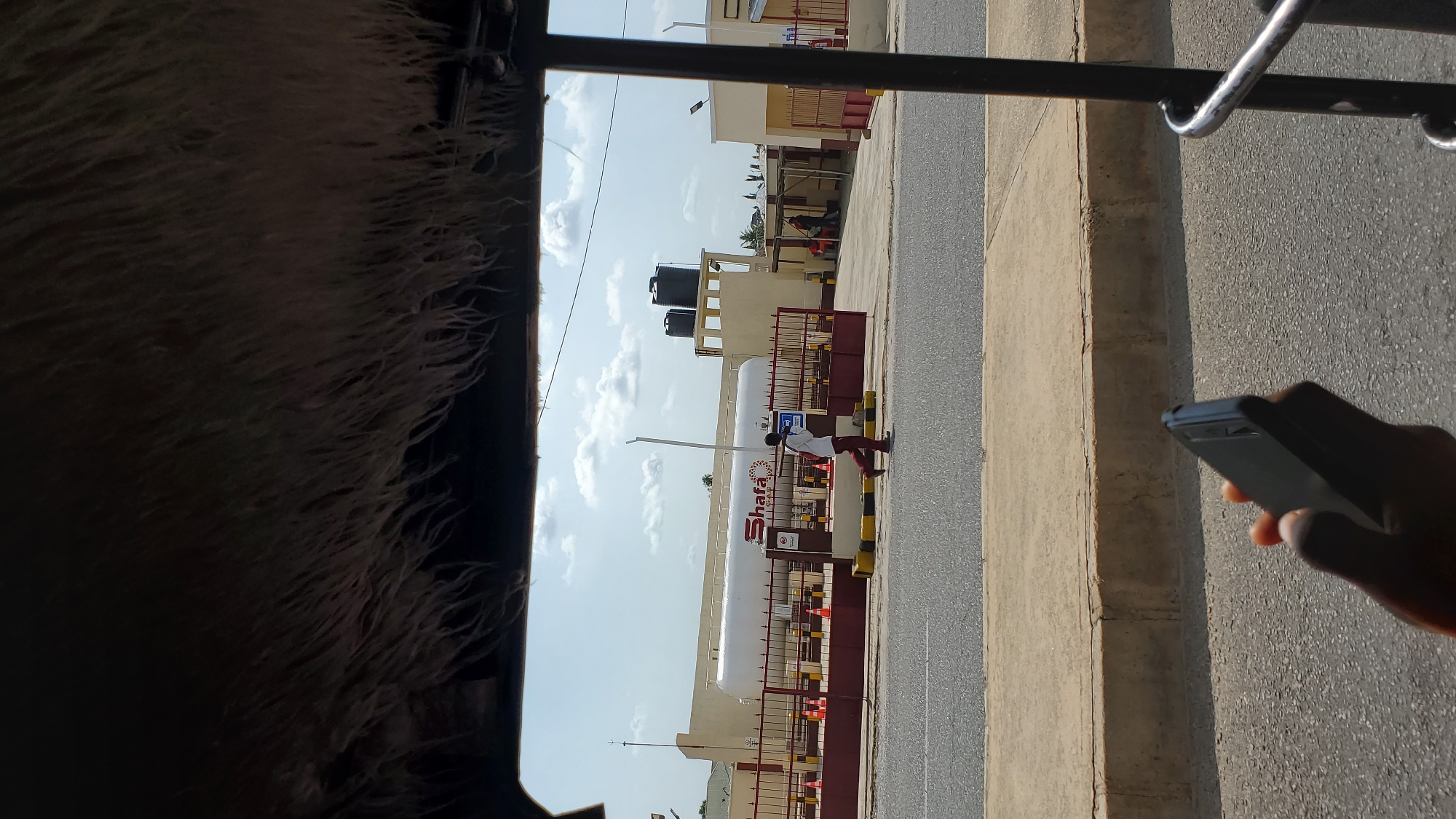 The image captures a street view in Jalingo, Nigeria, dominated by an industrial facility featuring a large white gas tank with "Shafa" branding. In the foreground, a person is walking across a paved road, and a hand holding a black smartphone is visible at the bottom of the frame. The industrial site is enclosed by a red metal fence with orange cones placed along it. The sky is partly cloudy. The perspective suggests the photo was taken from a vehicle.