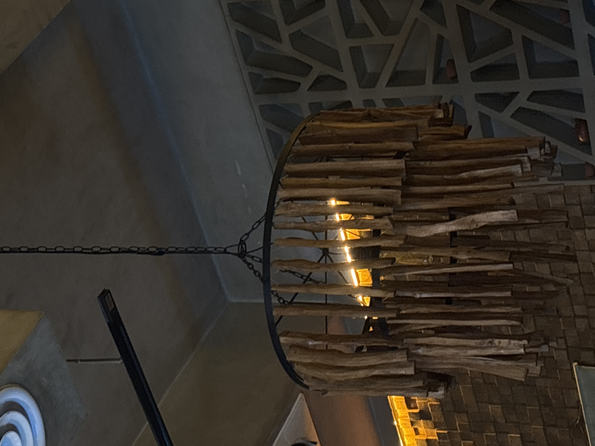 The image shows an ornate light fixture hanging from a ceiling. The fixture is made of a dark metal ring and horizontal pieces of light-colored wood. It is suspended by a chain. The ceiling above the fixture has a geometric design. The image was taken in North Kuta, Indonesia.