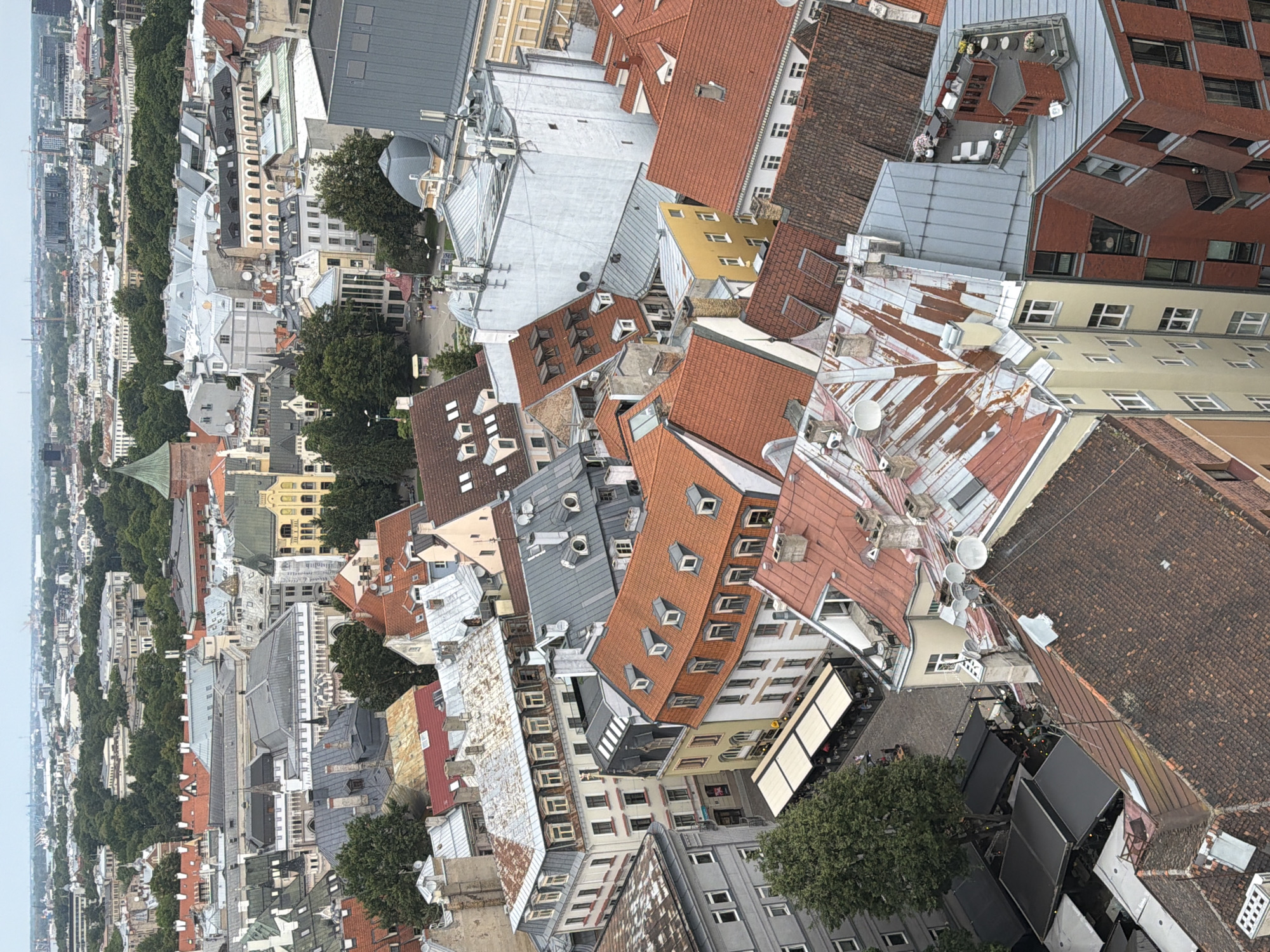 This image is an aerial view of the city of Riga, Latvia, focusing on a dense urban landscape characterized by historic architecture and a mix of building styles. The scene is dominated by rooftops, predominantly red-tiled and some made of metal, with varying states of repair. Many buildings feature dormer windows and chimneys.
Several prominent structures are visible. A large, light-colored building with many arched windows is situated towards the top left. To its right, a building with a distinctive green spire and a red-tiled roof stands out. In the center and foreground, a cluster of multi-story buildings with orange and red tiled roofs are packed closely together, showcasing a consistent architectural style that suggests an older part of the city. Some of these buildings have weathered metal roofs with visible seams and patching.
On the ground level, visible in the lower left quadrant, is a street scene with outdoor seating areas for what appear to be cafes or restaurants, partially covered by black and white awnings. A large green tree provides a touch of nature amidst the urban density. In the background, a wider city panorama unfolds, with more buildings, trees, and a hint of a body of water or industrial area in the distance under a cloudy sky. The overall impression is one of a well-established European city with a rich architectural heritage.