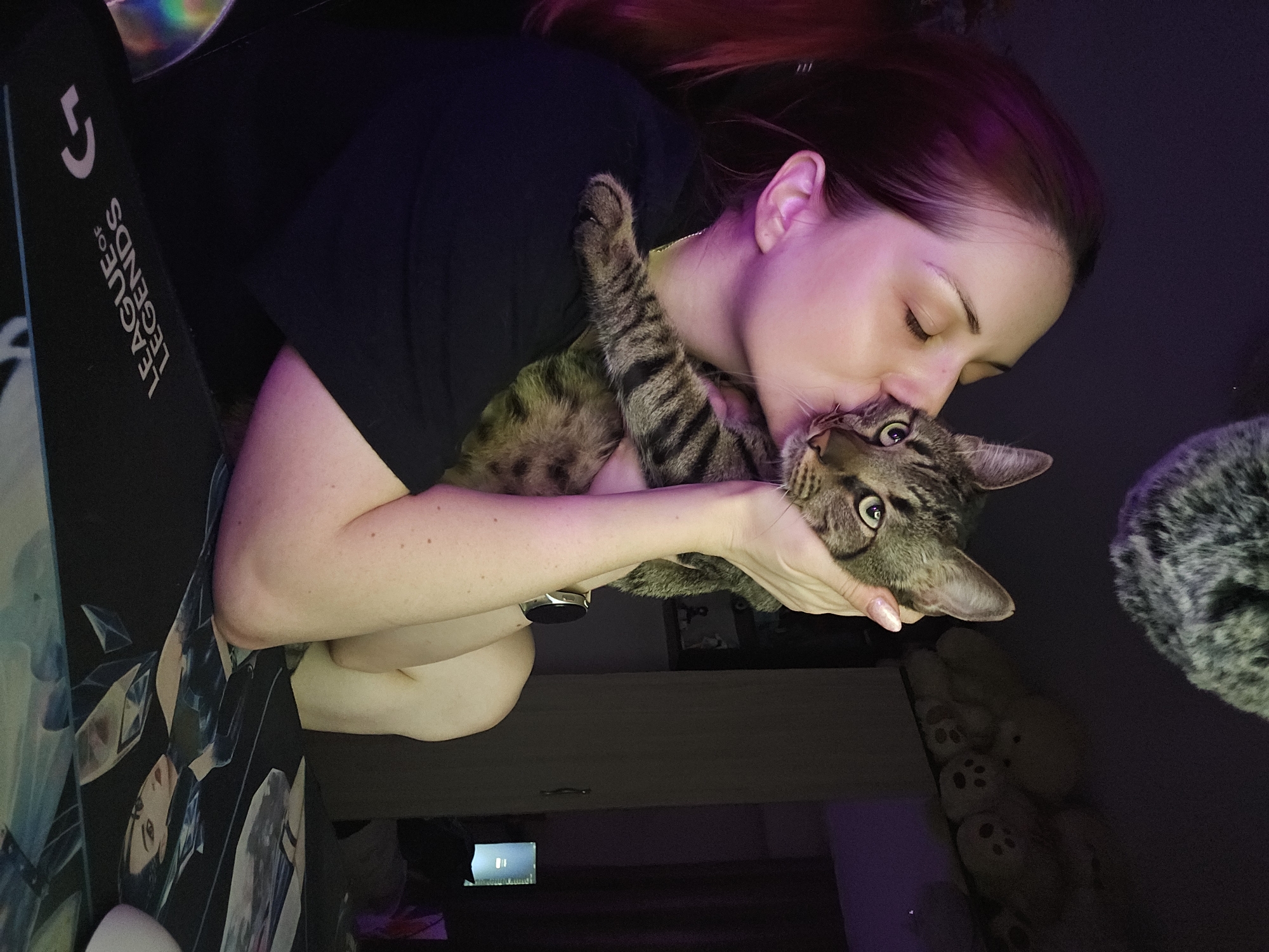 A young adult female with reddish-brown hair, wearing a dark short-sleeved top and a silver-toned smartwatch on her left wrist, is seated indoors under low, ambient purple and pink lighting. She is holding a brown and black striped tabby cat with both hands and is kissing its head, with her eyes closed in an affectionate gesture. The cat, with light green-yellow eyes, rests a front paw on her shoulder and looks calmly towards the viewer. In the foreground, a black desk mousepad is visible, displaying the inverted text "LEAGUE OF LEGENDS" and a Logitech "G" logo, alongside printed anime-style characters and a portion of a white computer mouse. An iridescent object is partially visible on the far left. In the background, a shelf holds several light-colored teddy bears with visible paw print details. To the right, dark furniture and a distant illuminated screen are present. A fuzzy grey object hangs in the upper left background. The scene is located in the city of Burgas, Bulgaria.