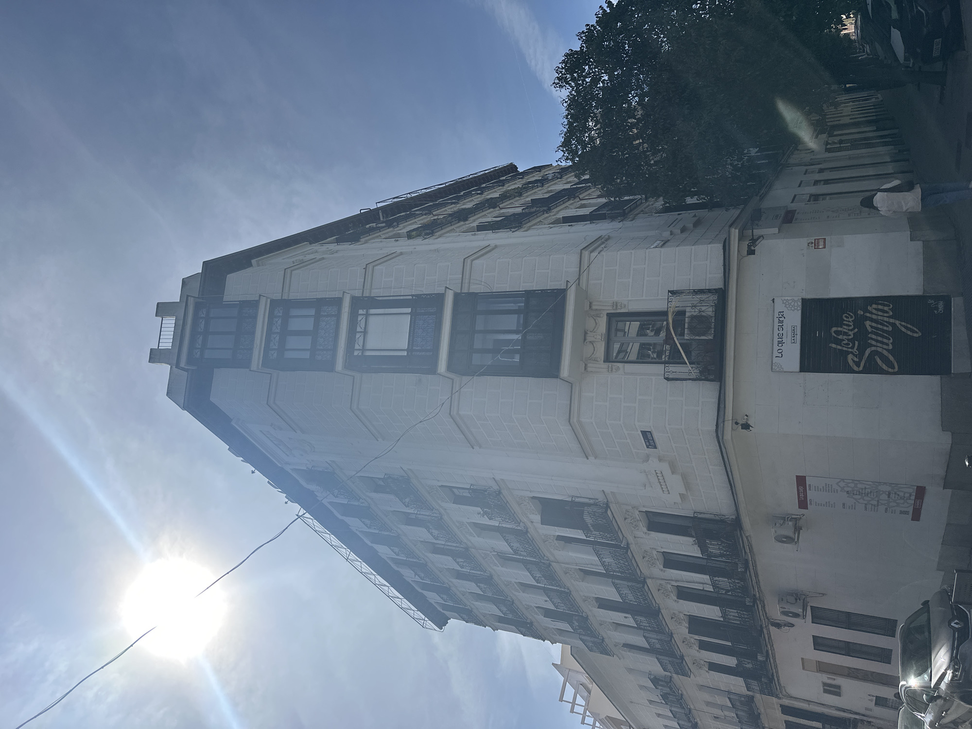 The image shows a multi-story building in Madrid, Spain. The building is light-colored with decorative architectural details, including balconies and window grilles. A sign on the corner of the building reads "Lo que Surja." The sky is visible in the background with the sun creating a strong glare. A tree is partially visible on the right side of the building. A person is walking on the sidewalk next to the building and parked cars are visible on the street.