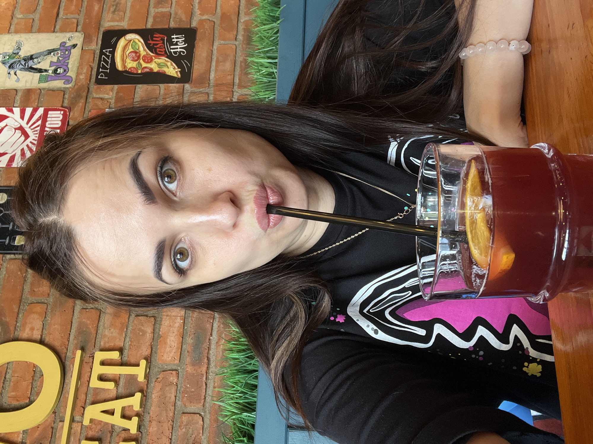 A young woman with long, dark hair drinks a beverage through a black straw in a restaurant, sitting at a wooden table. She wears a black t-shirt with a graphic design and a rose quartz bracelet. The drink is in a tall glass, with a visible orange slice. The background includes a brick wall adorned with novelty signs featuring images of a pizza, Joker, and fist. There is artificial grass along the top of the booth she is sitting in. A yellow sign is also visible on the brick wall.