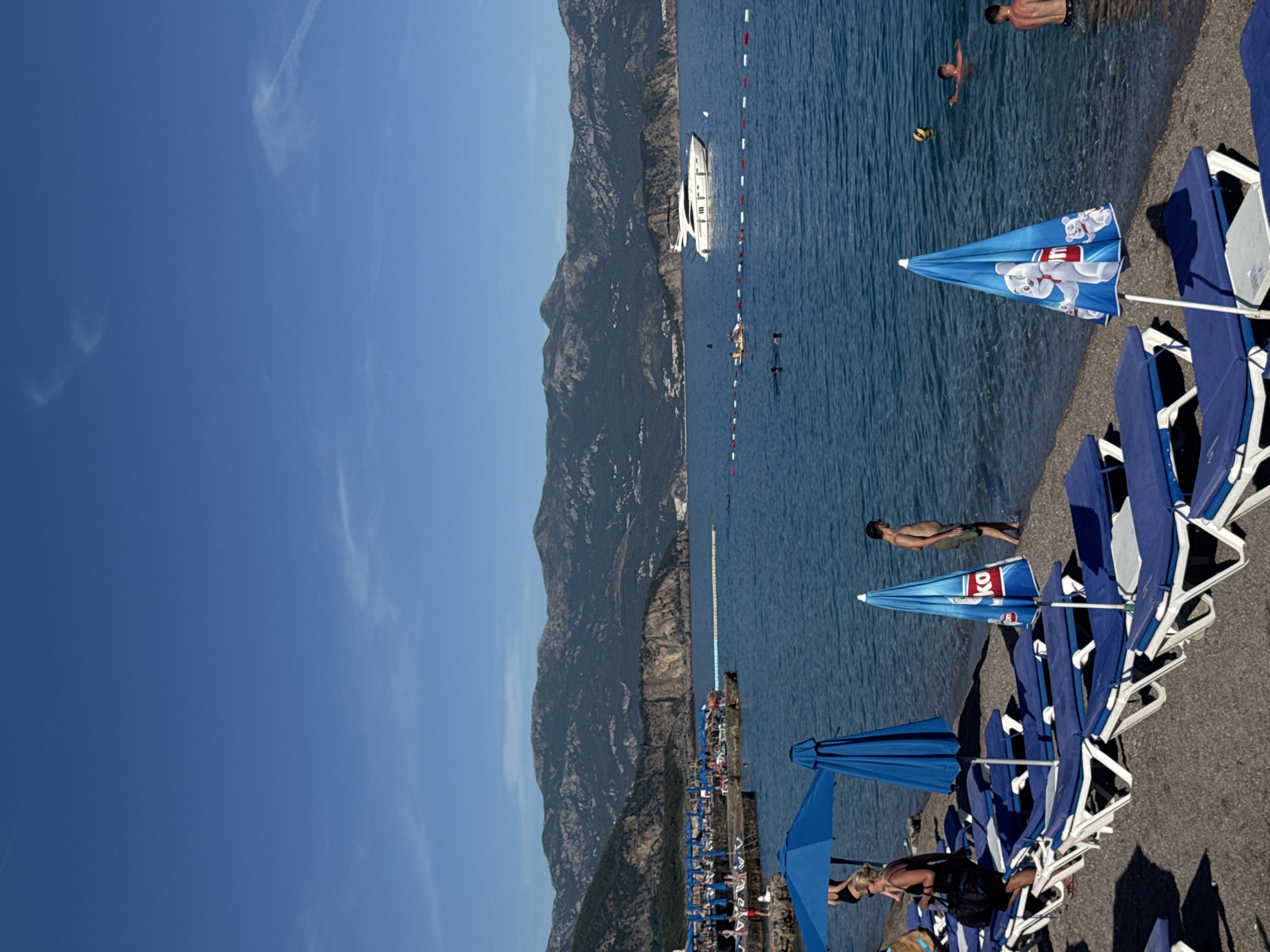 The image depicts a sunny day at a pebble beach in Prijevor, Montenegro. In the foreground, rows of blue lounge chairs with white frames are neatly arranged on the beach. Several blue umbrellas are set up, some providing shade. One umbrella in the middle ground features a cartoonish design of a bear. A woman in dark clothing is seen walking near the lounge chairs, carrying a black bag.
The sea is calm and blue, with several people swimming and enjoying the water. A white yacht is anchored in the distance, and a small raft with people on it is visible further out in the water. A red and white buoys line separates the swimming area.
The background is dominated by a large, rugged mountain range with sparse vegetation. The sky is a clear, bright blue with a few wispy white clouds. The overall scene conveys a relaxed atmosphere of a popular tourist destination on the Montenegrin coast.