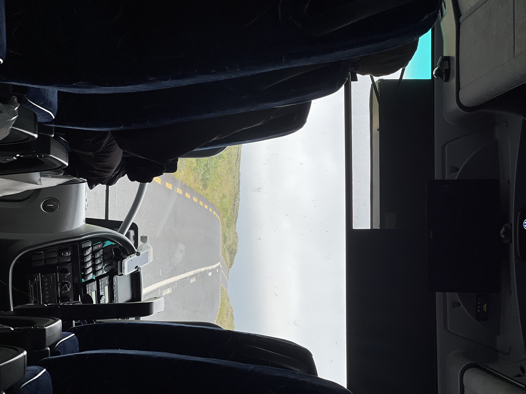 The image shows the view from inside a bus, looking forward through the windshield. The seats of the bus are in the foreground, with a person seated in the driver's position, partially obscured by the seats and dashboard. The road ahead is paved and has yellow dashed lines marking the lanes. The road winds through a grassy, hilly landscape under an overcast sky. The scene is consistent with the rural landscape of Meenacreevagh, Ireland.