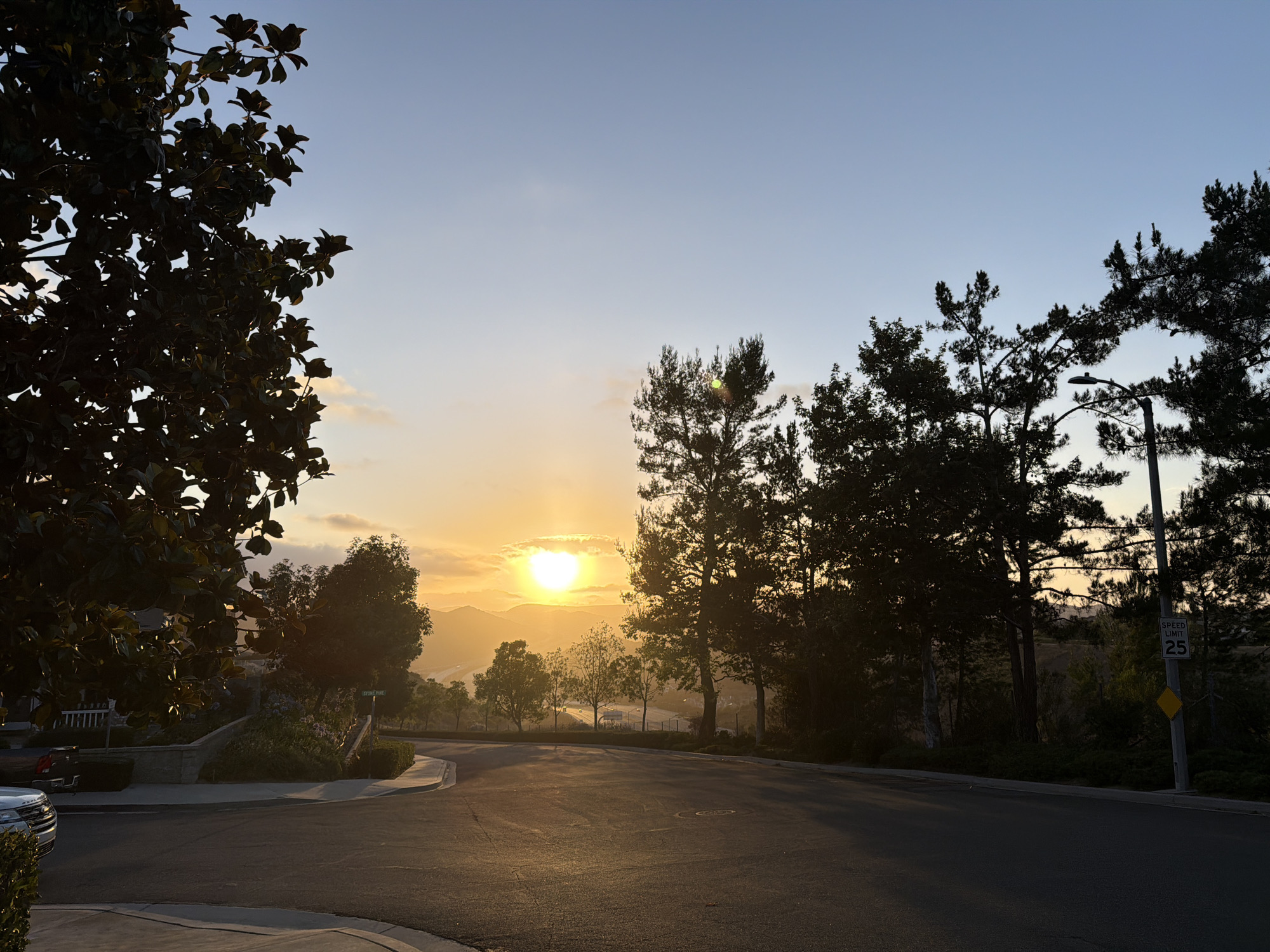 The image shows a street in Aliso Viejo, California, USA, during sunset. The sun is setting behind rolling hills in the distance, casting a warm glow over the landscape. Silhouetted trees line the street, and a speed limit sign reads "SPEED LIMIT 25". In the foreground, a paved road curves to the right, with a sidewalk and manicured landscaping on the left. The sky is a gradient of blue and orange, with a few wispy clouds. There are no people or animals visible in the image.