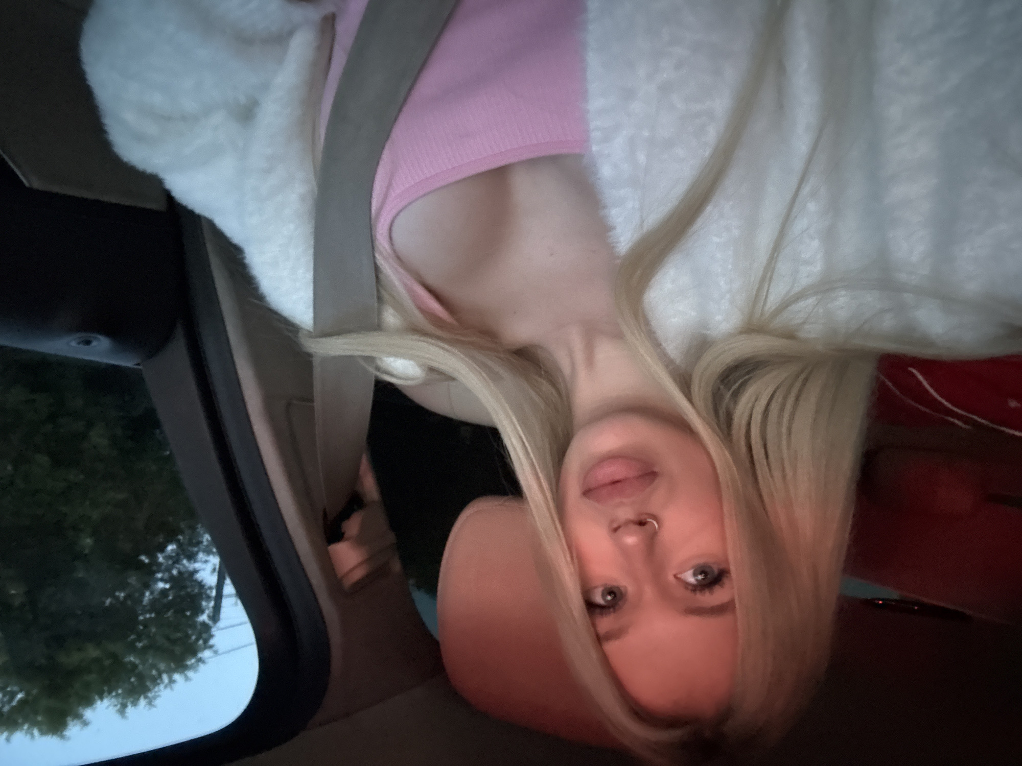 A young woman with blonde hair and blue eyes is sitting in the passenger seat of a car. She is wearing a pink tank top and a white fuzzy jacket. Her nose is pierced with a silver ring. A seatbelt is fastened across her chest. The interior of the car is visible, including a headrest and part of the dashboard. Through the car window, trees and the sky are visible, suggesting an outdoor environment, possibly on a road outside of Saint Petersburg, Russia, during twilight hours. The lighting is dim, with some red light sources reflected on the left side of the image.