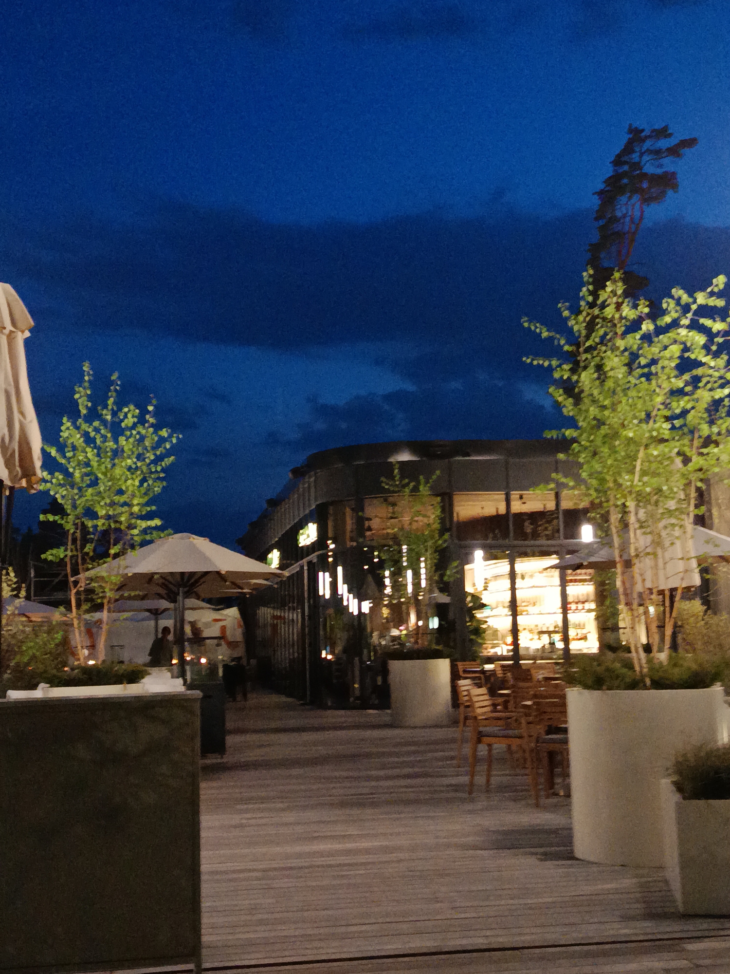 The image depicts an outdoor dining area at night. The location is Razdory, Russia. The area features wooden decking, with tables and chairs arranged near a building. There are trees in planters and umbrellas providing shade. The building's interior is illuminated. The sky is dark, with some clouds. A taller tree stands in the background on the right. There are a few people visible in the background, under the umbrellas.