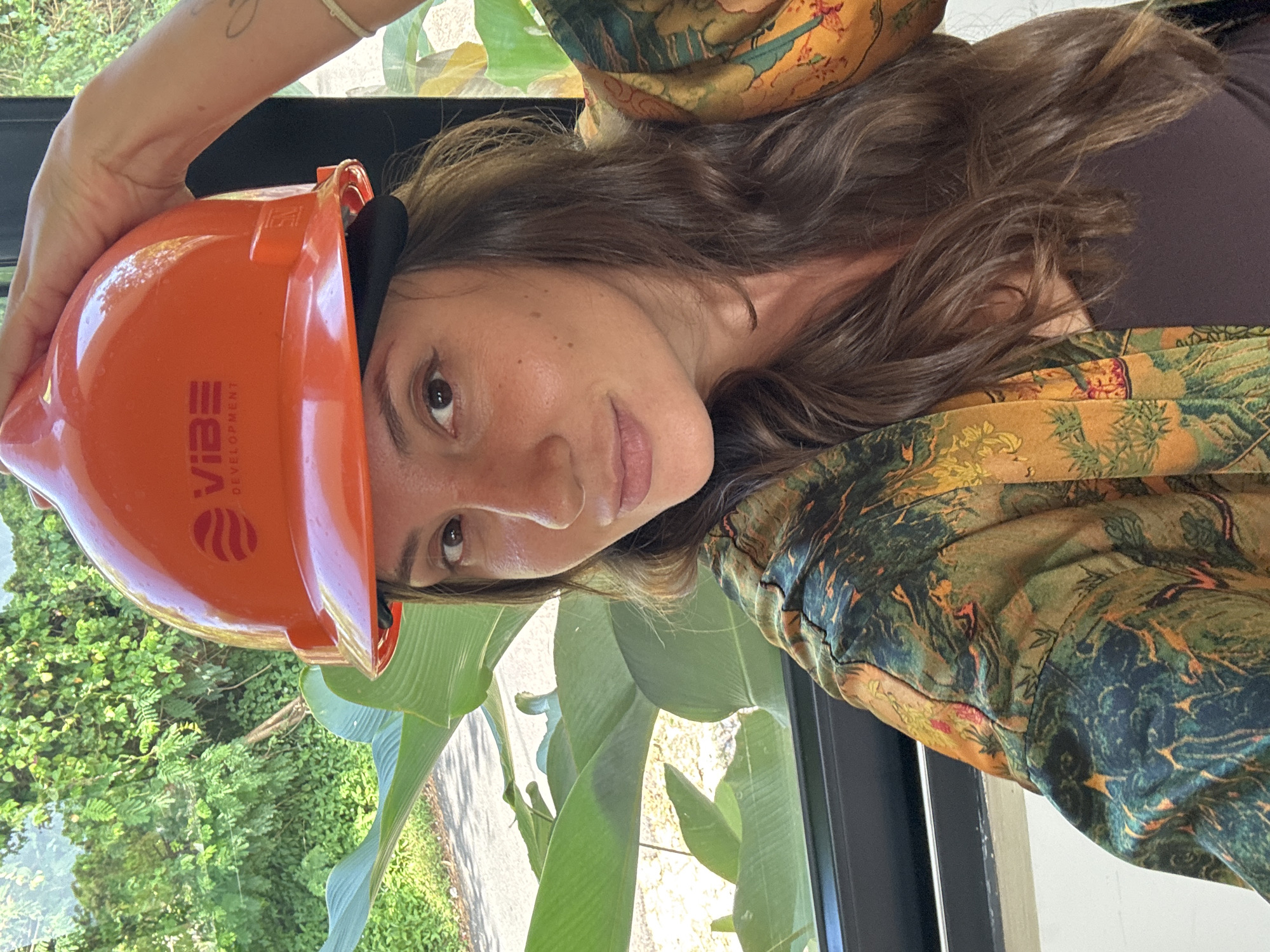 A woman is shown in a close-up, medium shot, holding an orange hard hat with the logo "VIBE DEVELOPMENT" on it. She has brown hair and is wearing a dark-colored top with a patterned, green and yellow jacket or shawl draped around her shoulders. The background shows a window with a view of greenery, suggesting an outdoor or garden setting. The image appears to have been taken indoors, close to the window. There is no information available to definitively place this image in Kuta Selatan, Indonesia.