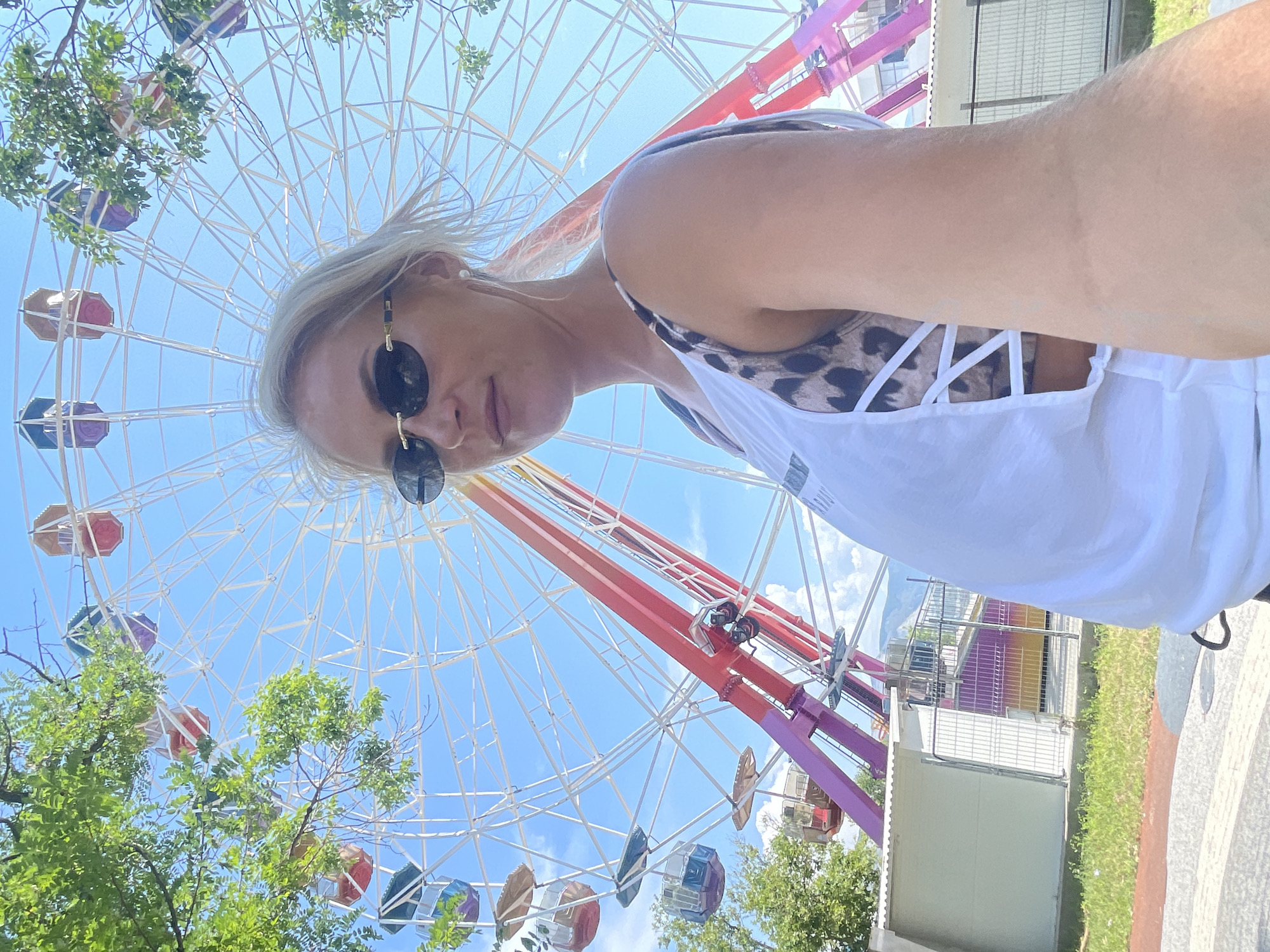 A woman is taking a selfie in front of a Ferris wheel at an amusement park. She is wearing sunglasses and a white top over a leopard-print bikini top. The Ferris wheel is large and colorful, with red, pink, and yellow cabins. Trees and a blue sky with clouds are visible in the background. The setting appears to be a sunny day in Novorossiysk, Russia.