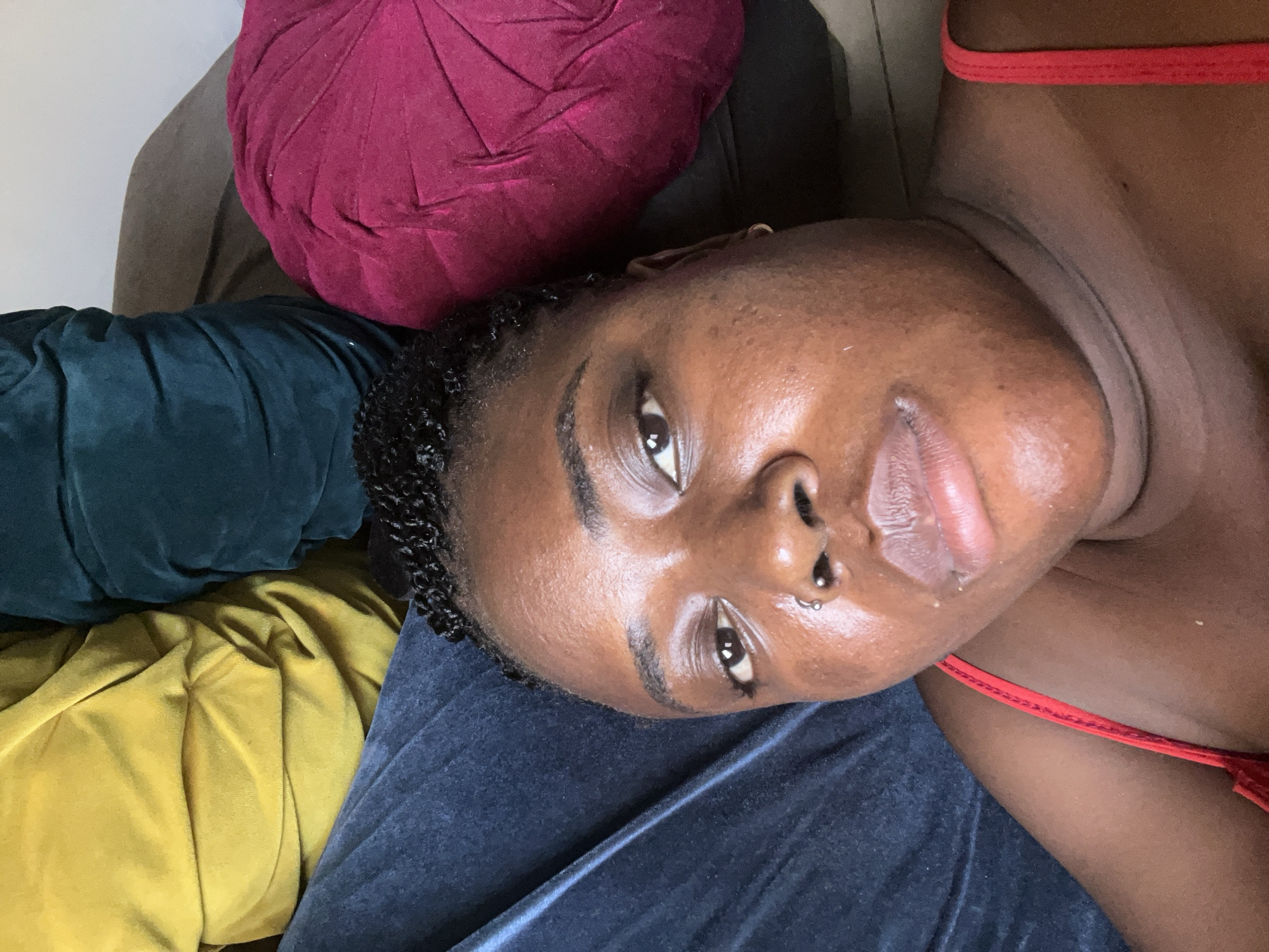 The image is a close-up selfie of a person lying down, with their face filling most of the frame. The background consists of several large, plush cushions in vibrant colors: yellow, teal, and fuchsia. The person has dark skin, dark braided hair pulled up, and is wearing a red top. They have a nose ring and a subtle earring. The lighting suggests an indoor setting, possibly a living room or bedroom. There is no specific indication of the city of Abuja, Nigeria, within the image itself. The focus is on the person and the colorful soft furnishings surrounding them.