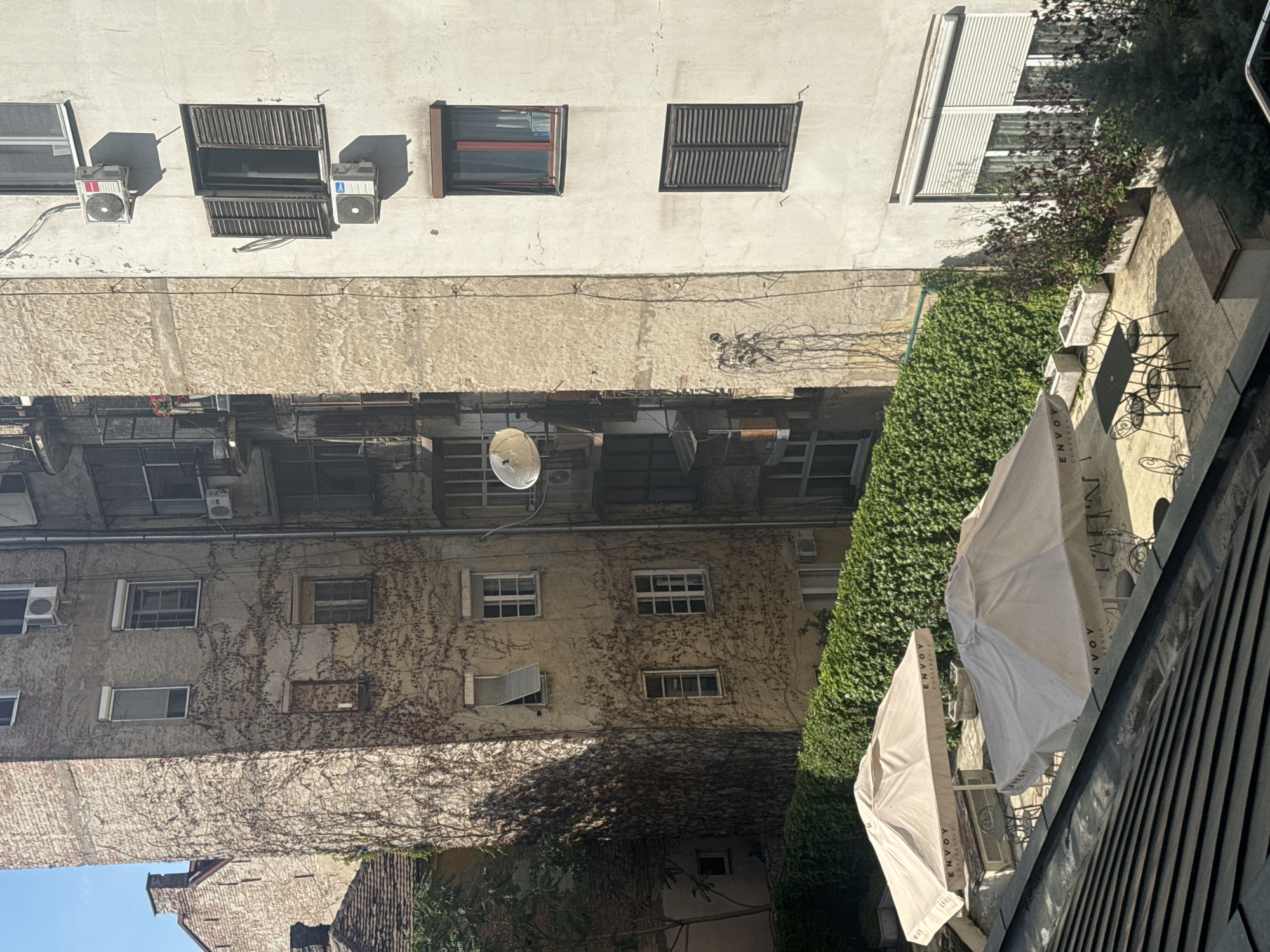 This is an exterior shot taken from a high vantage point, looking down and across at several buildings and outdoor spaces. The scene appears to be an urban courtyard or alleyway, with multiple stories of buildings visible. The architecture is varied, with some buildings featuring old plaster walls and exposed brick, while others have smoother, modern facades with white paint. Many windows are visible, some with shutters and some without. Several air conditioning units are mounted on the exterior walls of the buildings.
Vines and ivy cover large portions of the building exteriors, suggesting that this is a well-established urban area. In the foreground, there is a paved outdoor area with tables and chairs, set up as if for a cafe or restaurant. Two large, light-colored umbrellas are set up over the tables, providing shade. The umbrellas have text visible, including "ENVOY" and "SINCE 2014."
The lighting suggests it is daytime, with strong shadows cast by the buildings and objects. The overall atmosphere is one of a lived-in urban environment, possibly in a city with a Mediterranean or Balkan climate due to the greenery and outdoor cafe setup. The orientation of the photo is rotated 90 degrees counterclockwise, so the image appears sideways.
