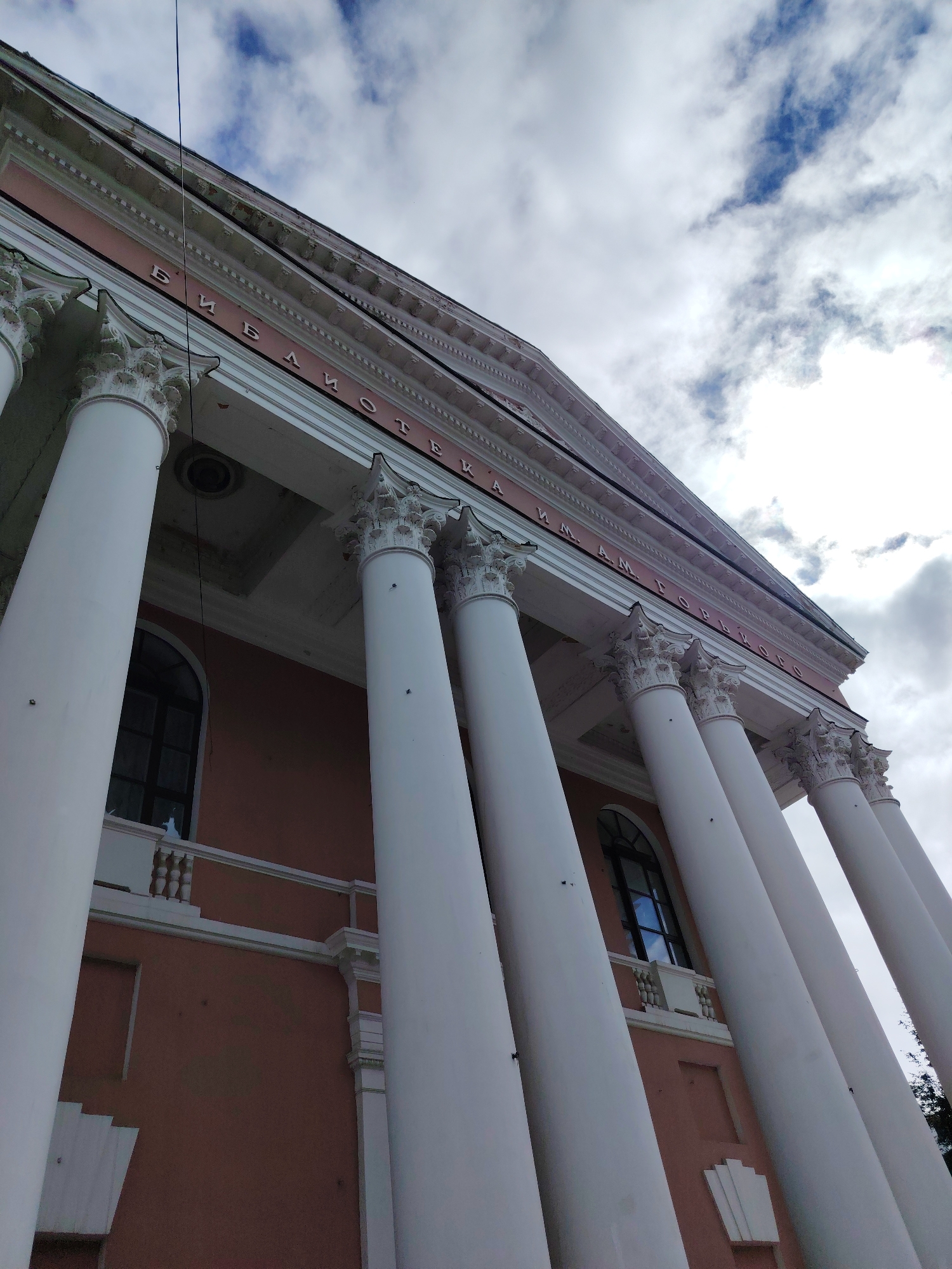 This is a low-angle, full-shot photograph of the Tver State Technical University Library, located in Tver, Russia. The facade of the building is dominated by a row of large, white Corinthian columns. The upper portion of the building features architectural lettering in Cyrillic script, reading "БИБЛИОТЕКА ИМ. А.М. ГОРЬКОГО," which translates to "Library named after A.M. Gorky." The building's walls are a muted pink color, and arched windows with dark frames are visible between the columns. The sky above is cloudy, with patches of blue sky peeking through. The overall impression is one of grandeur and classical architecture.