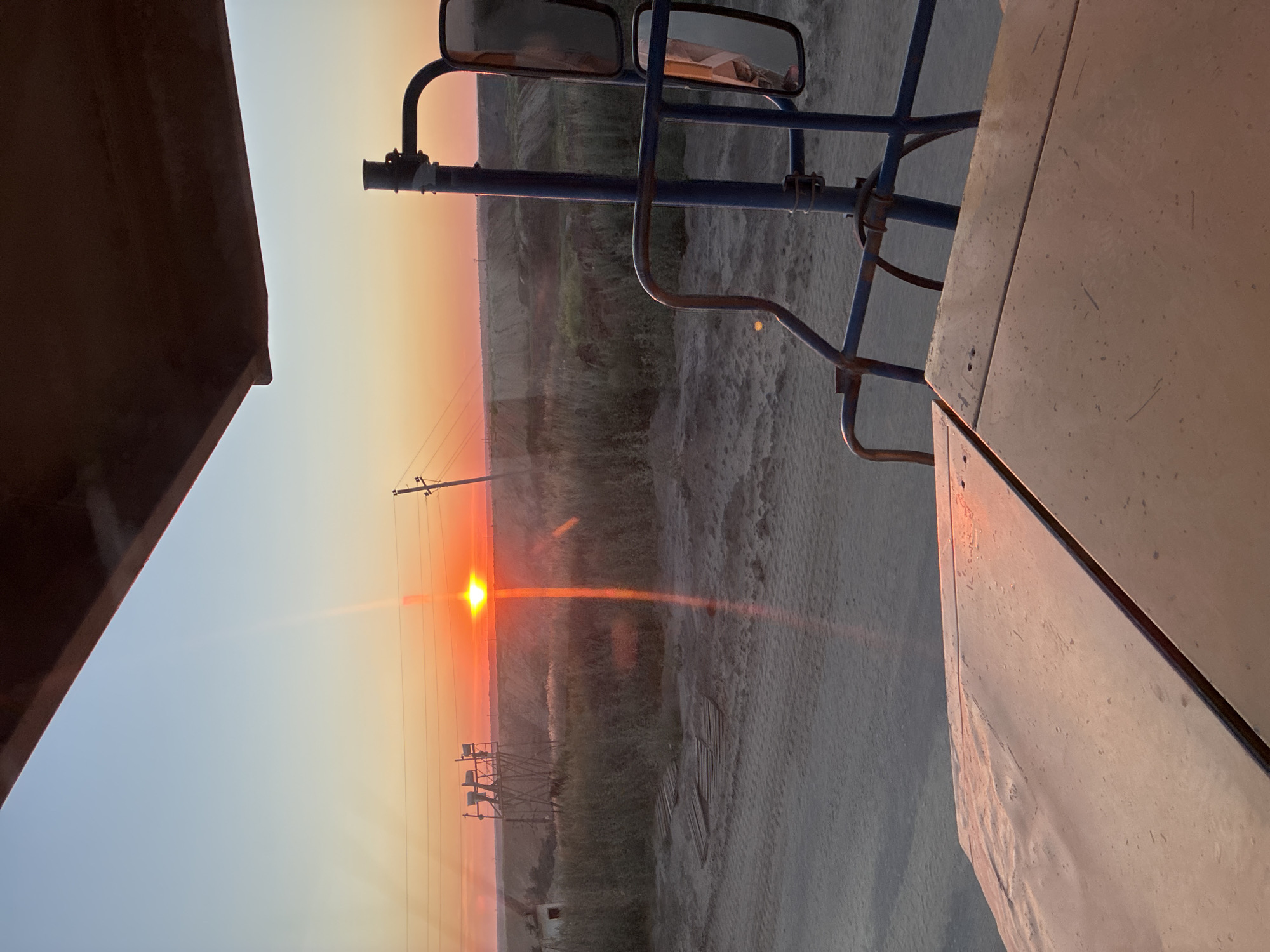 The image captures a sunset scene from the perspective of what appears to be a vehicle or piece of heavy machinery. The sun is a bright orange orb low on the horizon, casting a warm glow and creating a lens flare that extends upwards. The sky transitions from a pale blue at the top to vibrant orange and pink hues near the horizon. In the middle ground, there is a grassy, uneven terrain with some wooden planks scattered on the ground. Two wooden towers with electrical equipment are visible on the left, and utility poles with wires stretch across the landscape. The foreground is dominated by the metal surface of the machinery, which is reflecting the sunset's colors. A blue metal frame with mirrors is also visible, suggesting the vantage point is from a driver's cab or an operator's station. The overall environment appears to be an open, possibly industrial or rural area on the outskirts of Tatsinskaya, Russia, characterized by dusty ground and sparse vegetation.