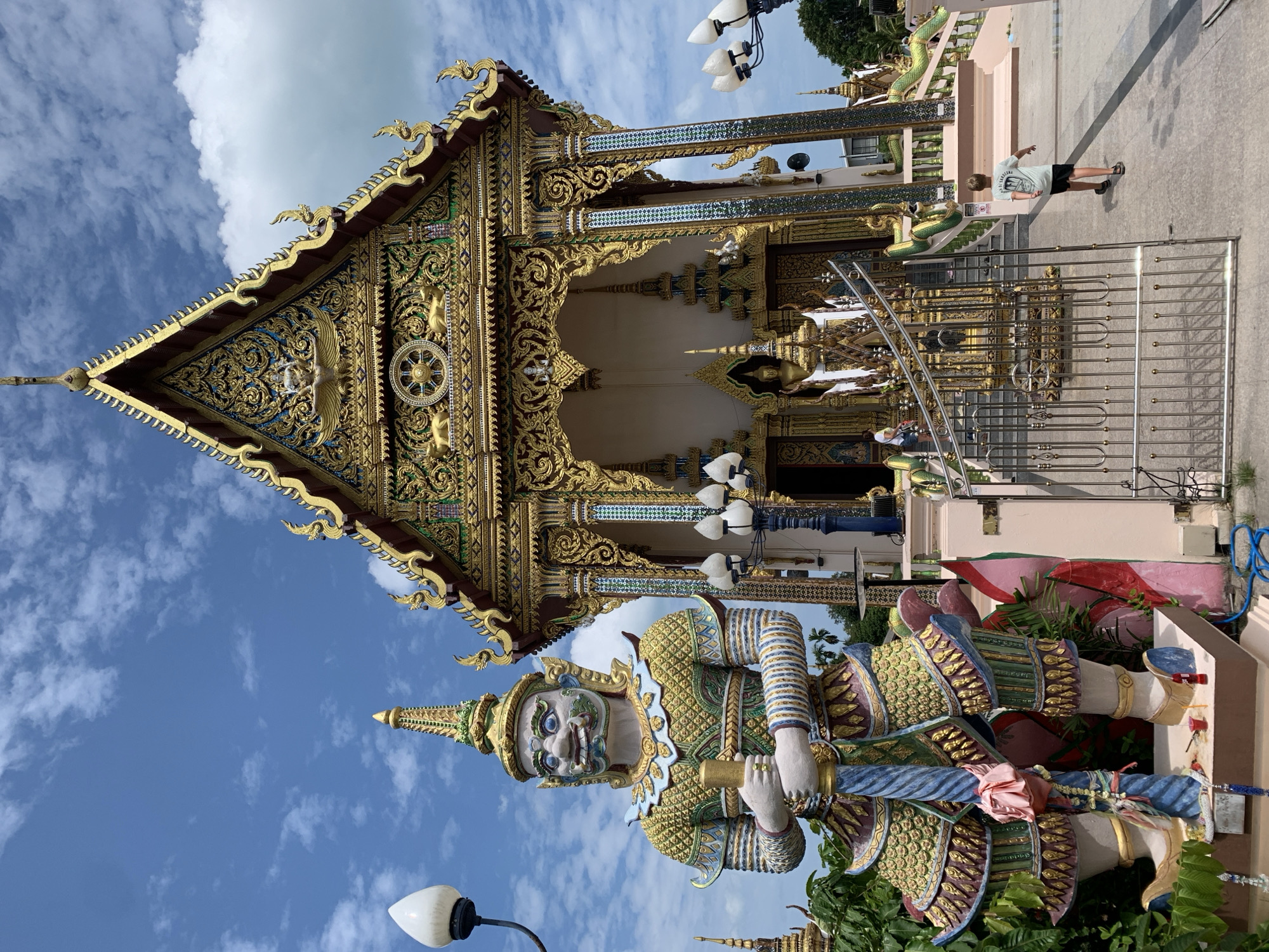 This image captures a scene at a temple in Ko Samui District, Thailand. In the foreground, a large, intricately decorated statue of a guardian figure stands prominently. The statue is dressed in ornate, colorful attire and holds a golden staff. To the right of the statue is the temple building, characterized by its tiered, golden roof adorned with elaborate carvings and decorative elements. A boy in a light-colored t-shirt and dark shorts is walking away from the camera towards the temple entrance. He is positioned on a paved area in front of a metal gate. The environment appears sunny with a blue sky dotted with white clouds. Lush green foliage is visible around the statue and on the left side of the temple. The overall atmosphere suggests a visit to a cultural and religious site.