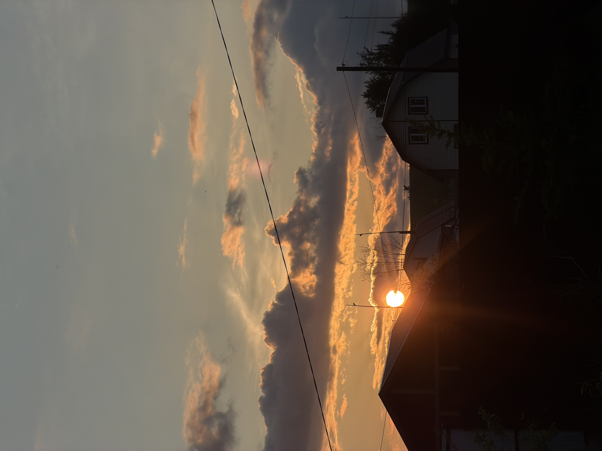 This is a vertically oriented, landscape-oriented photograph taken at sunset in Emel'yanovo, Russia. The scene depicts a rural or suburban setting with a focus on the dramatic sky.
The sky is filled with a mix of dark, brooding clouds and wispy, illuminated clouds tinged with orange and pink hues, indicative of the setting sun. A single, bright streetlamp is visible, its light a prominent focal point in the middle right of the frame, casting a warm glow. The sun itself is a bright, intense orb peeking through the clouds, its rays emanating outwards.
In the background, two houses are partially visible, silhouetted against the colorful sky. The house on the left appears to be a lighter color with visible windows, while the structure on the right is almost entirely in shadow. Power lines are strung across the upper portion of the image, adding to the sense of a lived-in, developed landscape. The overall mood is serene and somewhat melancholic, characterized by the fading light and the quiet presence of the houses. The time of day is clearly sunset, and the weather appears to be clear with dramatic cloud formations. No visible text is present in the image.