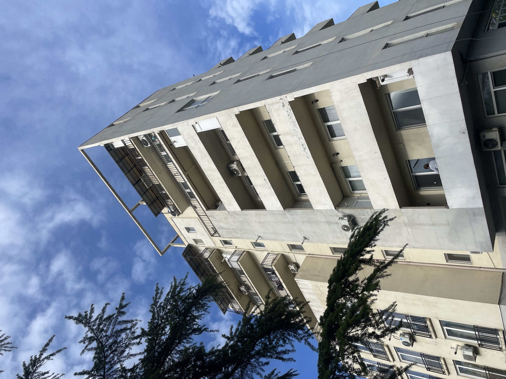 A low-angle shot shows a tall, multi-story building in Tbilisi, Georgia, with a blue sky and scattered clouds in the background. The building is light gray with white accents and has numerous windows and balconies. Air conditioning units are visible on the exterior walls. Two evergreen trees partially obscure the lower portion of the building.