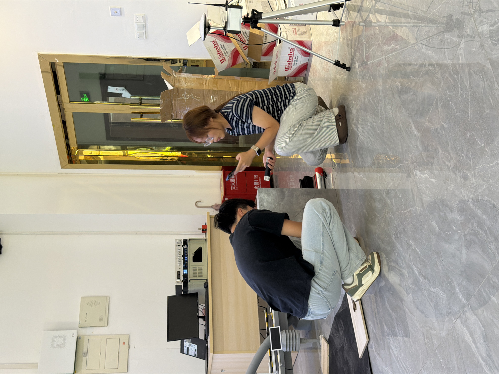 The image depicts two individuals, a male and a female, crouching on a polished gray tiled floor in what appears to be an indoor laboratory or workshop. The male, wearing a black t-shirt and light blue jeans, is facing away from the camera, tending to a large metallic rectangular object on the floor. The female, with glasses and her hair tied back, is facing the male and holding a black device, possibly a remote control or a measuring tool, in her left hand. She is wearing a striped t-shirt and light-colored pants.
The environment is cluttered with various objects. To the left, a wooden desk is visible with a laptop, monitor, and electronic equipment. Against the wall on the left, several white panels and a cream-colored square panel are mounted. In the background, a doorway with gold-colored trim leads to another room, with an "EXIT" sign illuminated in green. Cardboard boxes, some labeled with "Wahaha," are stacked near the doorway. A tripod with a camera or sensor is positioned to the right of the female. A red fire extinguisher is also visible on the far left, indicating a safety measure. The overall impression is of a functional space where some kind of assembly, testing, or research is taking place. The specific location in Jiangjin, China is not directly ascertainable from the visual cues alone.