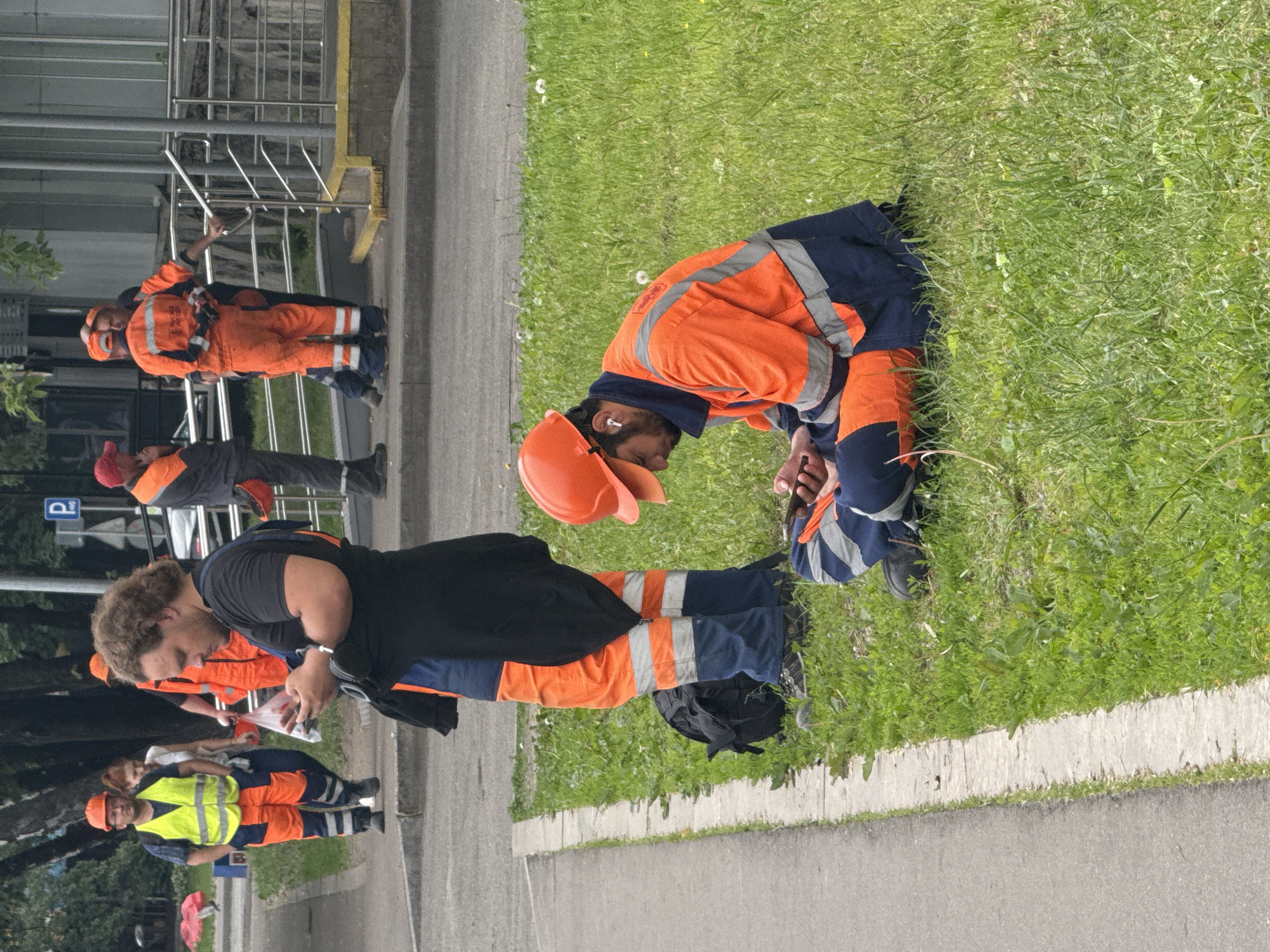 In Moscow, Russia, a group of workers in bright orange and blue uniforms are visible. Two men are standing and looking at their phones. One worker, wearing a yellow safety vest over his uniform, stands with his hands in his pockets, while another worker stands nearby. In the foreground, a man in an orange hard hat and uniform is sitting on the grass, looking at his phone. Another worker is also visible in the background, talking to a colleague. The environment appears to be a street or sidewalk next to a grassy area, with some trees and a building in the background. The overall scene depicts a moment of pause and rest for the construction workers.