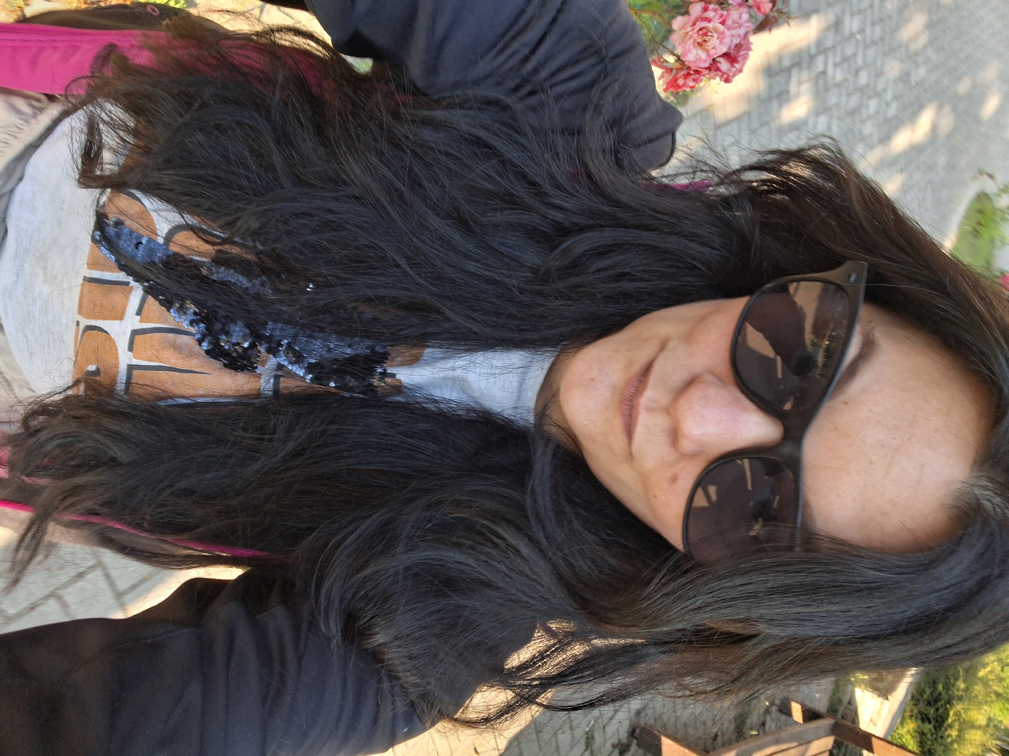 A medium shot selfie features a woman with long, dark, wavy hair and dark sunglasses. She is wearing a black jacket over a light-colored t-shirt with a graphic print and some sequined embellishments. Her hair is partially obscuring her face. In the background, a paved walkway, a wooden bench, and pink flowers are visible. The scene suggests an outdoor setting, possibly a park or garden. The sunlight indicates it is daytime. The background appears to be a paved area with cobblestone or similar material, common in older European city centers. The presence of a bench suggests a public space. The overall lighting indicates a sunny day. The woman is facing the camera, taking a selfie. The focus is on the woman, with the background slightly blurred.