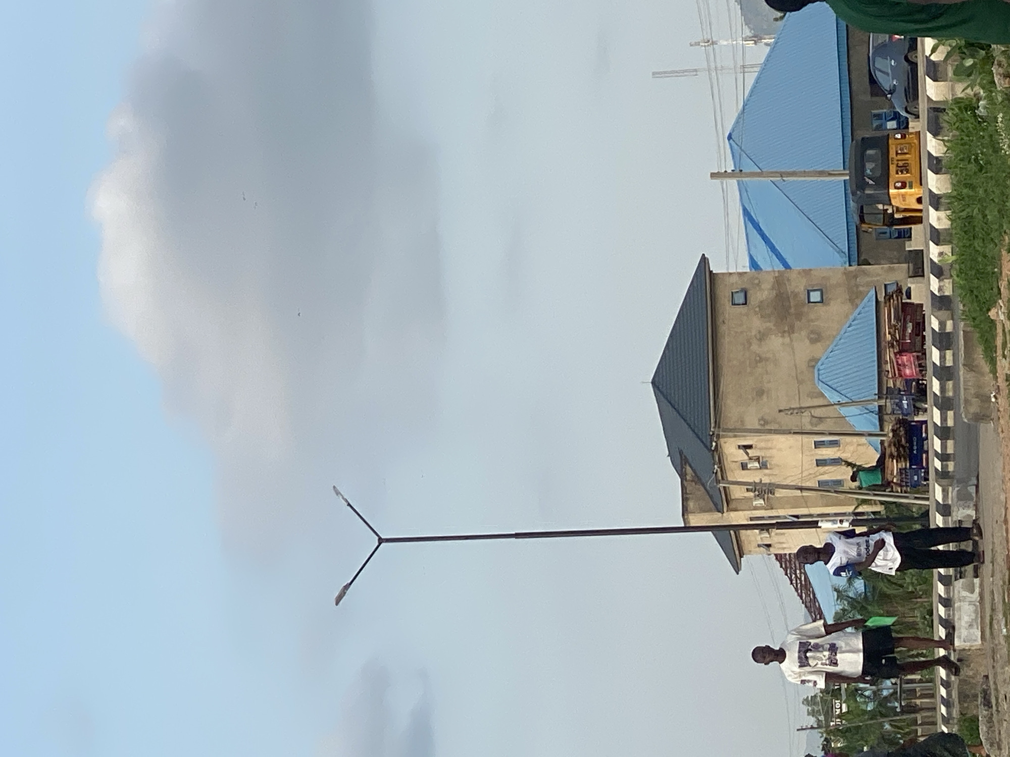 In Jalingo, Nigeria, two young men are standing on a concrete median strip beside a road. The man on the left, wearing a white t-shirt with a graphic and black shorts, holds a green folder. The man on the right, in a white t-shirt with blue trim and black pants, stands looking forward. Behind them, a tall street lamp with two lights extends upwards. Several buildings are visible, including a light brown, multi-story building with a dark blue roof, and another building with a bright blue roof. A yellow auto-rickshaw with the number "361" is parked on the road. A black car is also present. The sky is partly cloudy. The scene appears to be an urban or semi-urban environment.