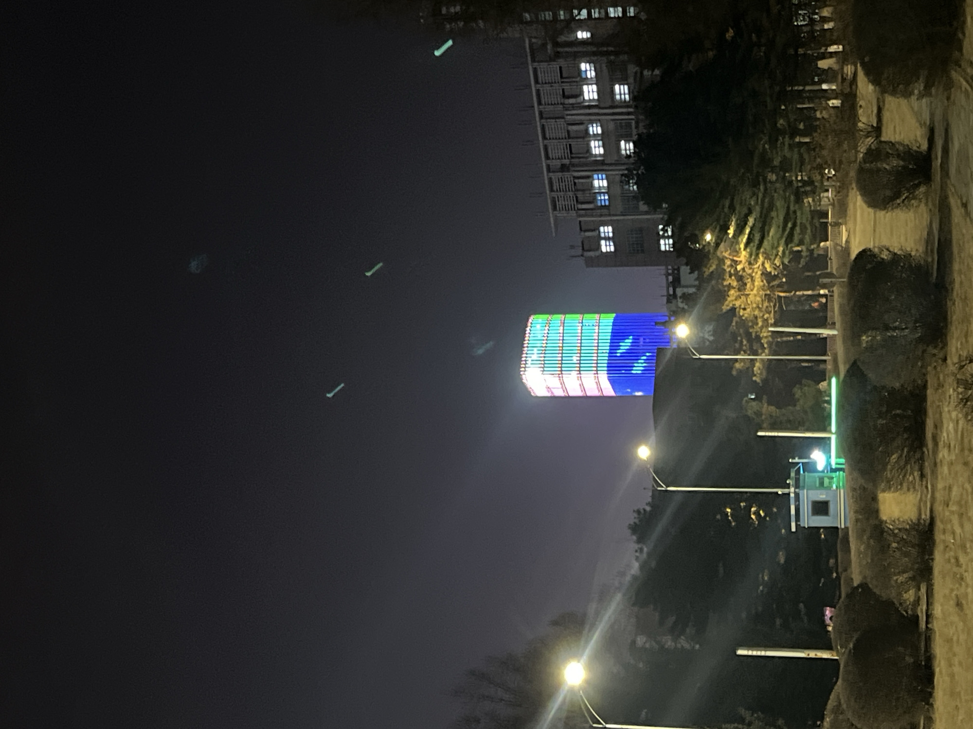 The image depicts a nighttime scene in Changchun City, Jilin Province, China, specifically on the campus of Northeast Normal University (东北师范大学). The central feature is a prominent, tall, rectangular building, identifiable as a main teaching or administrative building, whose entire facade is illuminated with dynamic LED lighting. This lighting displays a vertical gradient, transitioning from deep blue at the base, through lighter blue and green, to a pale green/white towards the top.
To the right of this LED-illuminated building, a large, multi-story building with a more conventional architectural style is visible. Its numerous windows glow with interior white light, and its upper levels feature distinctive horizontal architectural elements.
The foreground consists of dry, muted-colored ground, with several dark, rounded bushes and scattered evergreen and deciduous trees. Three streetlights with bright, warm yellow-white illumination are positioned in the mid-ground, casting light and producing visible lens flares. A small, light blue and white booth-like structure with a single window is located near the center-left foreground, partially illuminated by a streetlight and emitting a subtle green glow from its base. The dark sky above contains several horizontal and diagonal light green-blue streaks, indicative of lens flares or atmospheric effects. No individuals or vehicles are discernible in the scene.