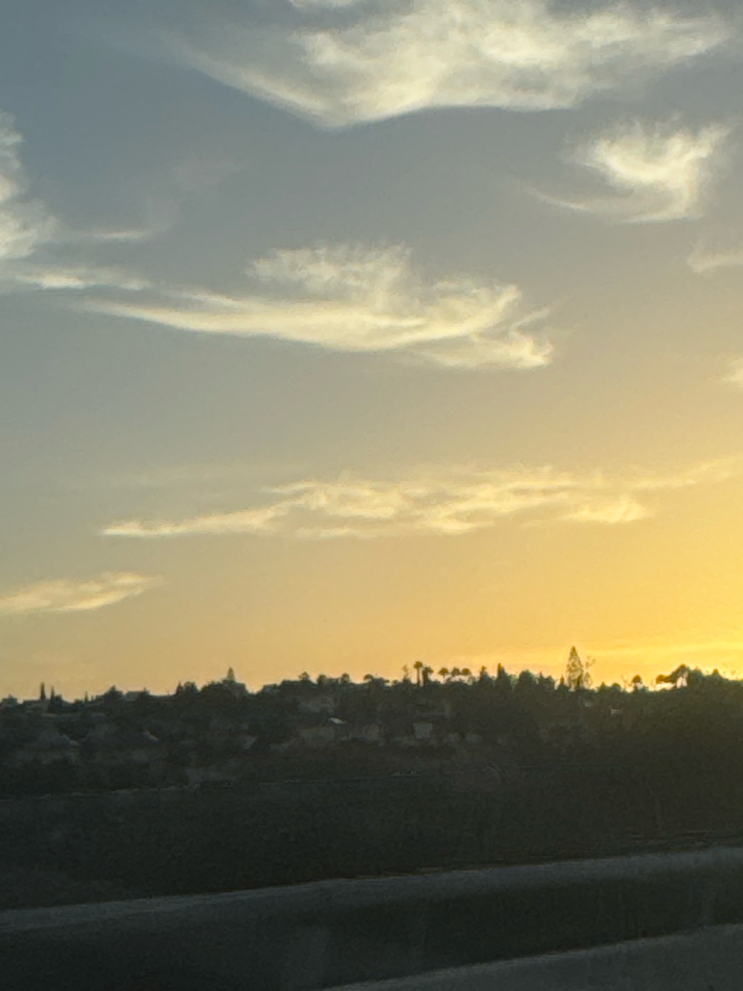 A landscape scene features a gradient sky transitioning from a pale blue to a vibrant yellow-orange near the horizon. Scattered white clouds are visible in the blue portion of the sky, with wispy, thin clouds also present. The horizon is marked by a silhouetted treeline and buildings, suggesting a settlement on a gentle slope. The foreground is dominated by dark, blurred shapes, possibly indicating a road or embankment. The image appears to be taken during a sunset or sunrise, given the warm colors of the sky. The location is identified as Germasogeia Municipality, Cyprus.