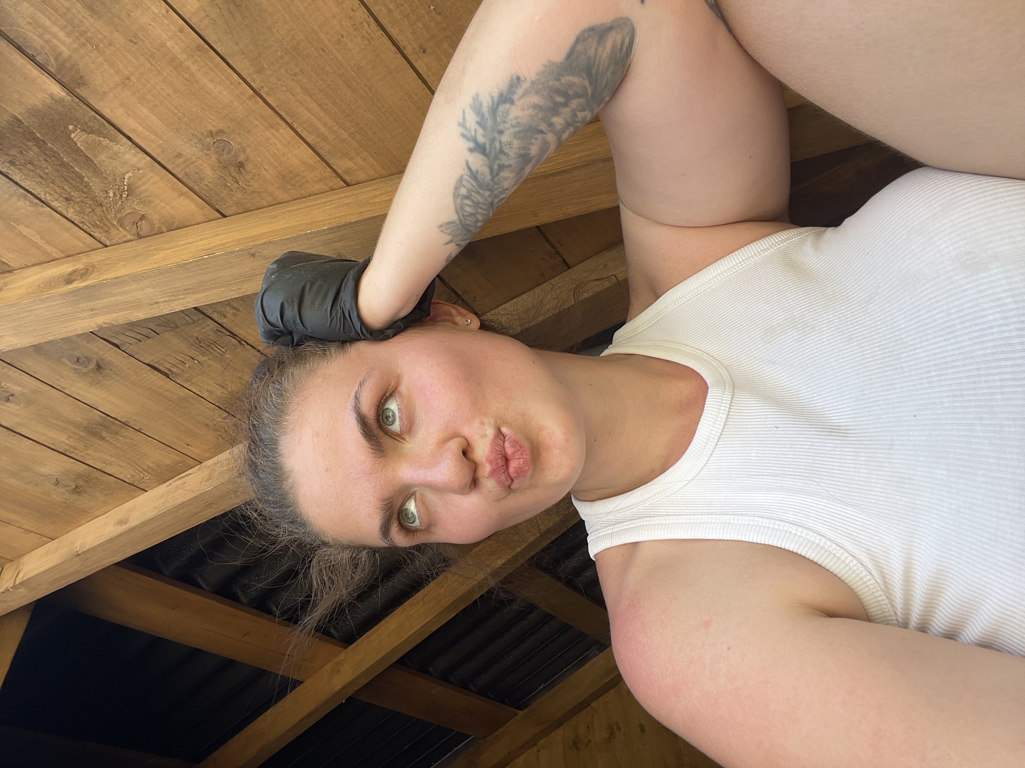 A woman with fair skin, green eyes, and light brown hair is pictured in a close-up, slightly tilted shot. She is wearing a white ribbed tank top and a black glove is visible on her right hand. A tattoo is visible on her right arm. Her lips are pursed in a pouting gesture. The background appears to be the interior of a wooden structure, possibly a shed or outbuilding. The structure appears to be located in the city of Мосметаллоконструкция Онт, Russia. The scene is lit with natural light.