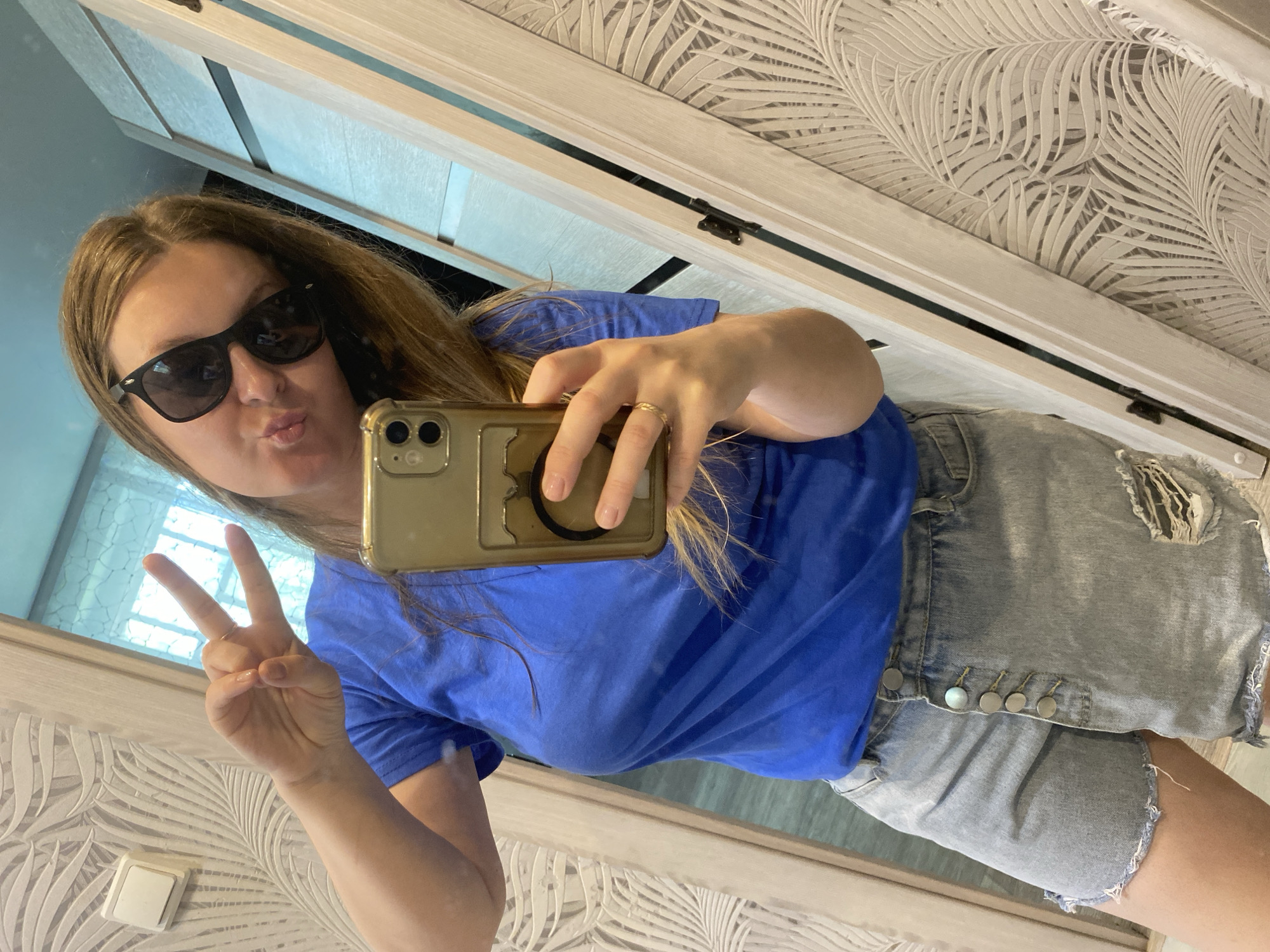 The image is a selfie taken by a woman in what appears to be an indoor environment. She is wearing sunglasses, a blue t-shirt, and denim shorts. She is holding a smartphone up to take the picture, while making a peace sign with her left hand and pursing her lips. The background shows a decorated wall with a white and grey leaf pattern, and a partially visible window to the left. There's also part of a light-colored door or wall frame visible. There is no specific information or details indicating that the location is in the city of Noviy, Russia.