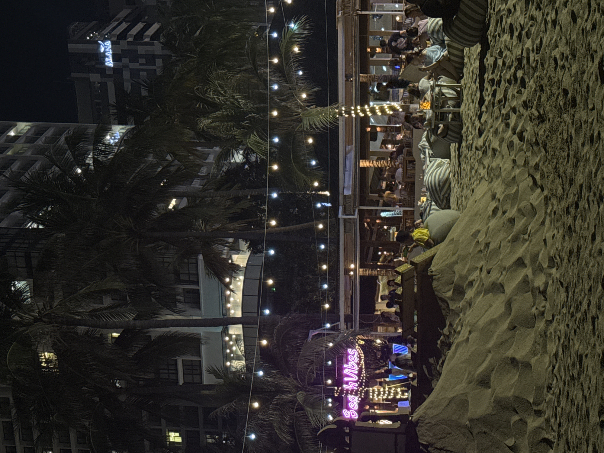 The image depicts an outdoor dining establishment situated on a beach in Nha Trang Ward, Vietnam, during nighttime. The restaurant appears to be crowded with people dining. Overhead string lights illuminate the area, adding to the ambiance. In the foreground, there is a sandy beach and in the background, there is a multi-story hotel with the word "ALANA" prominently displayed near the top. A neon sign with the words "Beach Vibes" is visible near the lower portion of the restaurant area. Palm trees are interspersed throughout the scene, contributing to the tropical atmosphere.