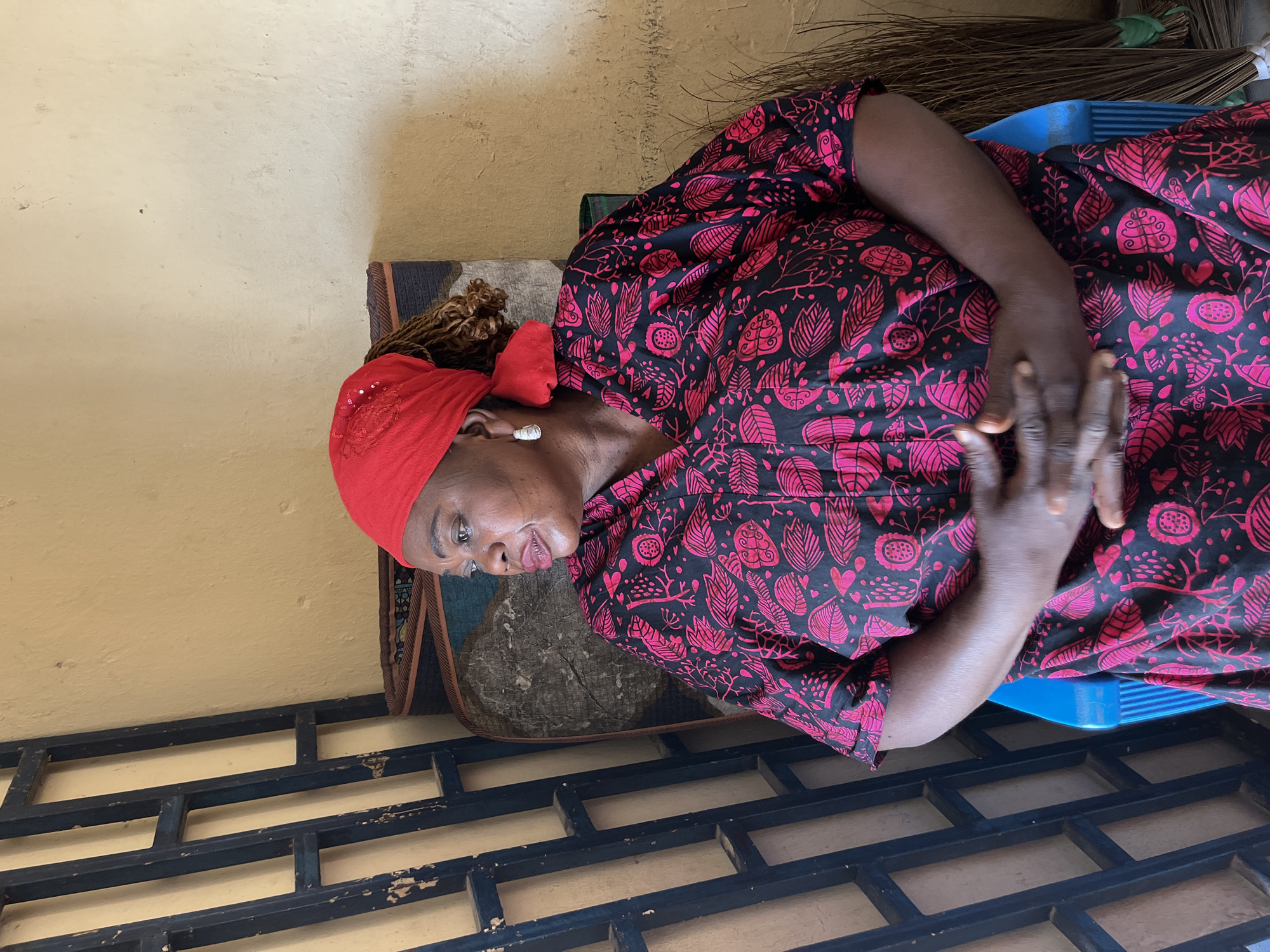 An adult woman is pictured seated in what appears to be an indoor or semi-outdoor setting in Jos, Nigeria. She has dark skin and is wearing a black dress featuring a vibrant pattern of pink leaves and floral designs. A bright red headwrap covers her hair, from which brown dreadlocks or braids are visible. She also wears large white stud earrings. Her hands are clasped together in front of her, resting on her lap.
The woman is sitting on a blue plastic chair, parts of which are visible under her and behind her. A patterned fabric or cushion can also be seen on the chair. The background consists of a light tan or beige wall with a visible textured finish. In the upper right section of the image, a bundle of thin, dry sticks, resembling brooms, leans against the wall. The foreground reveals a dark, gridded metal structure, possibly a window grille or a decorative barrier. The natural light suggests it is daytime. The woman's expression is calm and neutral, as she looks slightly to her left. No text is visible in the image. The scene conveys a moment of quiet repose in an everyday environment.