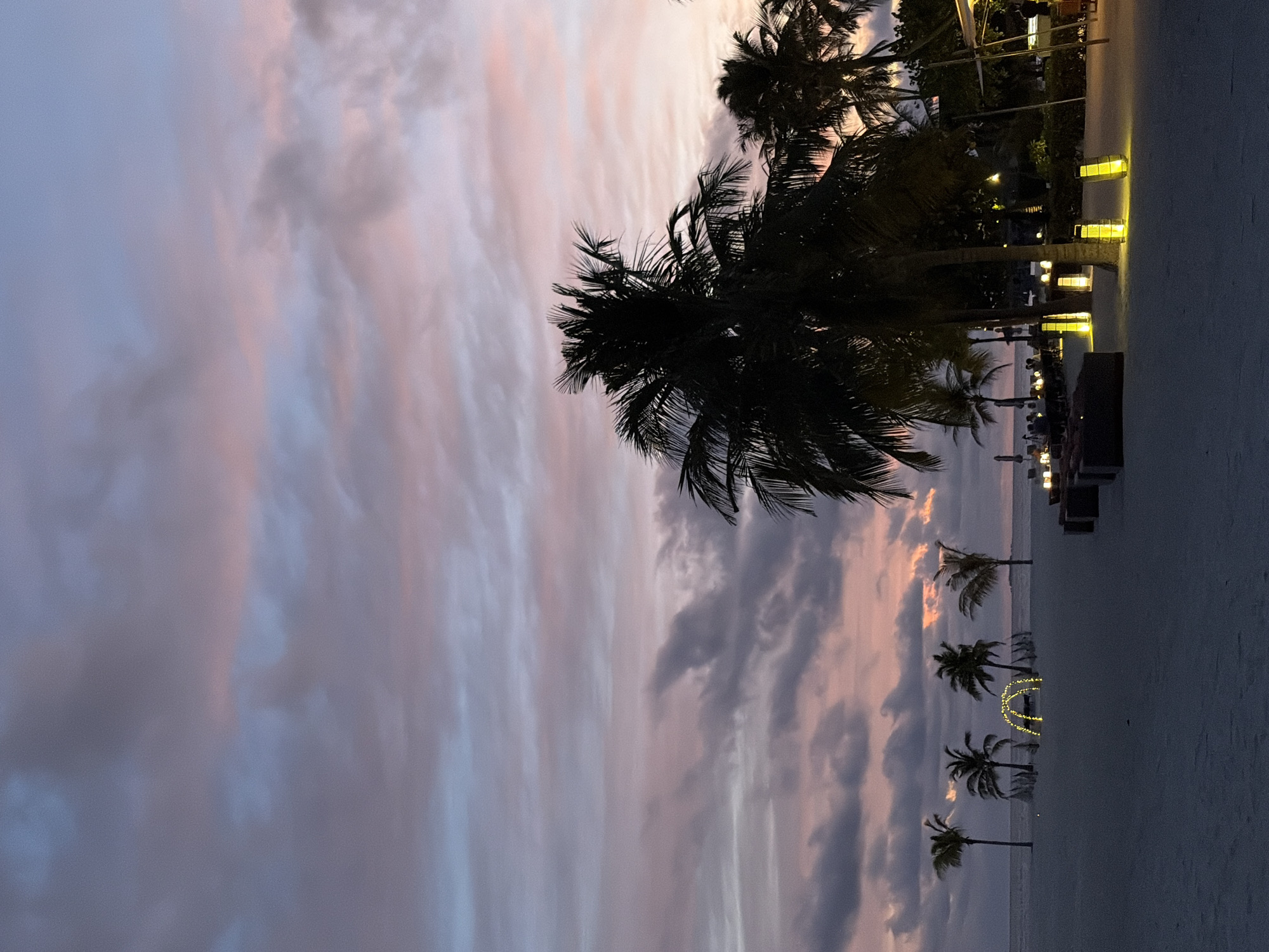 A serene beach scene at dusk in Halaveli, Maldives, features a wide expanse of white sand leading to a tranquil ocean. Several palm trees are silhouetted against a sky painted with soft hues of pink, purple, and blue from the setting sun. The scene is further enhanced by the warm glow of strategically placed yellow lanterns, illuminating the sandy pathway and casting soft light on the scattered lounge furniture. In the distance, a charming archway adorned with string lights adds a whimsical touch to the tropical setting. The overall ambiance is one of peaceful beauty and quietude, characteristic of a Maldivian sunset.