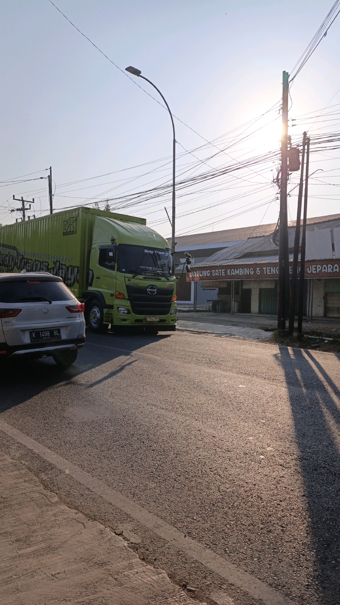 The image captures a bright, sunny day scene on a street in Kuwasen, Indonesia. A bright green truck with a large cargo container is prominently featured in the foreground, alongside the rear of a silver SUV. The truck has "Dedy Trans Jaya" text on its front windshield and a black license plate that reads "B 9541 WRX". The SUV has a white license plate that reads "K 1498 BL". In the background, a building with a sign that says "WARUNG SATE KAMBING & TENGE IN JEPARA" is visible. Utility poles with numerous wires are scattered throughout the scene, with the sun shining brightly through them, casting long shadows on the asphalt road. The road itself is textured with visible markings. The sky is clear and blue.
