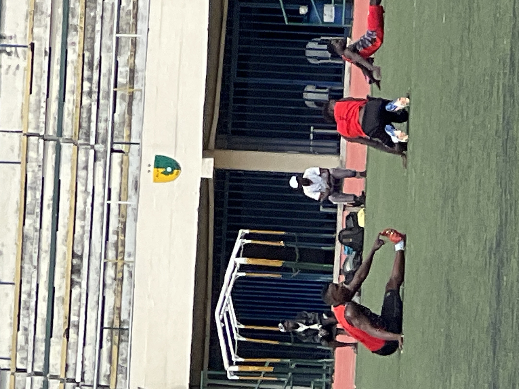 The image depicts a football training session at a stadium in Rafin Par, Nigeria. In the foreground, three young men in athletic wear are engaged in stretching exercises on a green artificial turf field. One man, wearing a red sleeveless shirt and black shorts, is stretching his left leg out in front of him, reaching for his toes. Another man, also in a red sleeveless shirt and black shorts, is on his knees, leaning forward. A third man, in a red and black patterned athletic shirt and red shorts, is stretching on the ground, with his body angled. In the background, several individuals are seated on a bench, observing the training. Behind them, a covered structure with yellow and white poles and a blue-barred fence is visible. Further in the background, stadium seating is present, with a crest featuring green and yellow colors displayed prominently. The overall scene suggests an active sports environment under daylight conditions.
