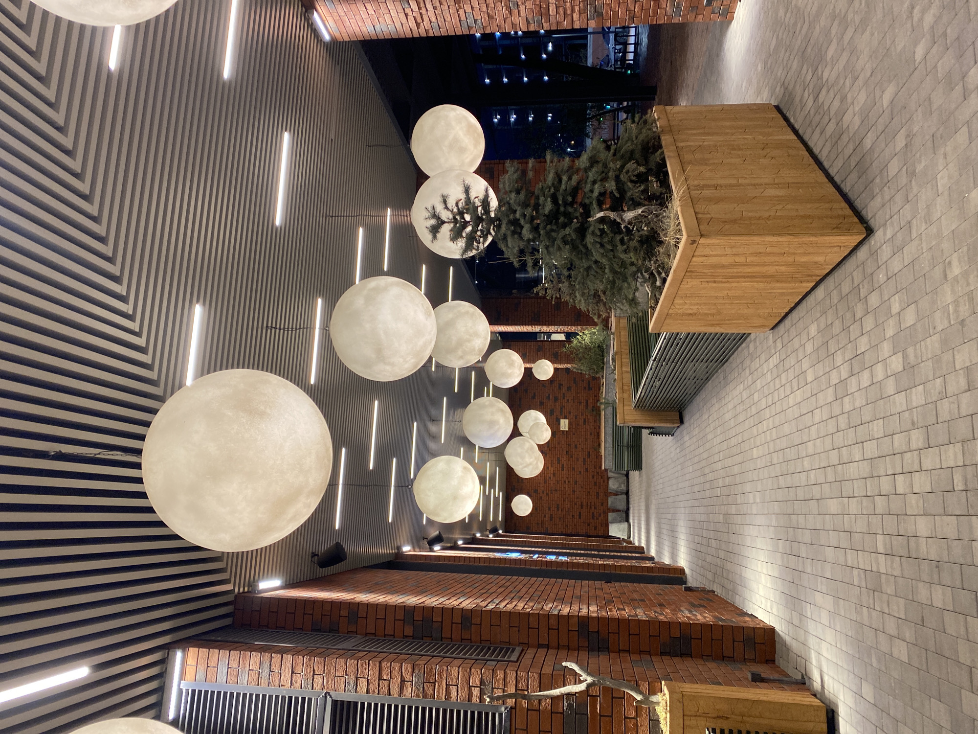 The image, originally rotated ninety degrees clockwise, presents a modern outdoor passage or courtyard viewed from a low angle, looking along its length. The overhead structure features a distinctive ceiling composed of parallel dark grey slats, with some sections forming a geometric chevron pattern. Numerous vertical strip lights are integrated into the ceiling, emitting a bright white light.
Dominating the upper space are multiple spherical light fixtures, varying in size, suspended from the ceiling. These luminaires glow with a soft, warm white light, strongly resembling full moons and adding a celestial element to the setting.
To the left, a wall constructed from dark reddish-brown bricks rises, while the ground-level wall on the right is made of lighter grey rectangular bricks. The ground itself is paved with similar light grey rectangular pavers. Along the right side, several large, angular planters crafted from light-colored wood house dark green evergreen trees or bushes, interspersed with some dried grasses.
The scene is set at night or late evening, as indicated by the darkness and the reliance on artificial lighting. No specific activities or people are visible, lending a serene and contemplative atmosphere to this modern urban space. No text is visible in the image. The location is Yekaterinburg, Russia.