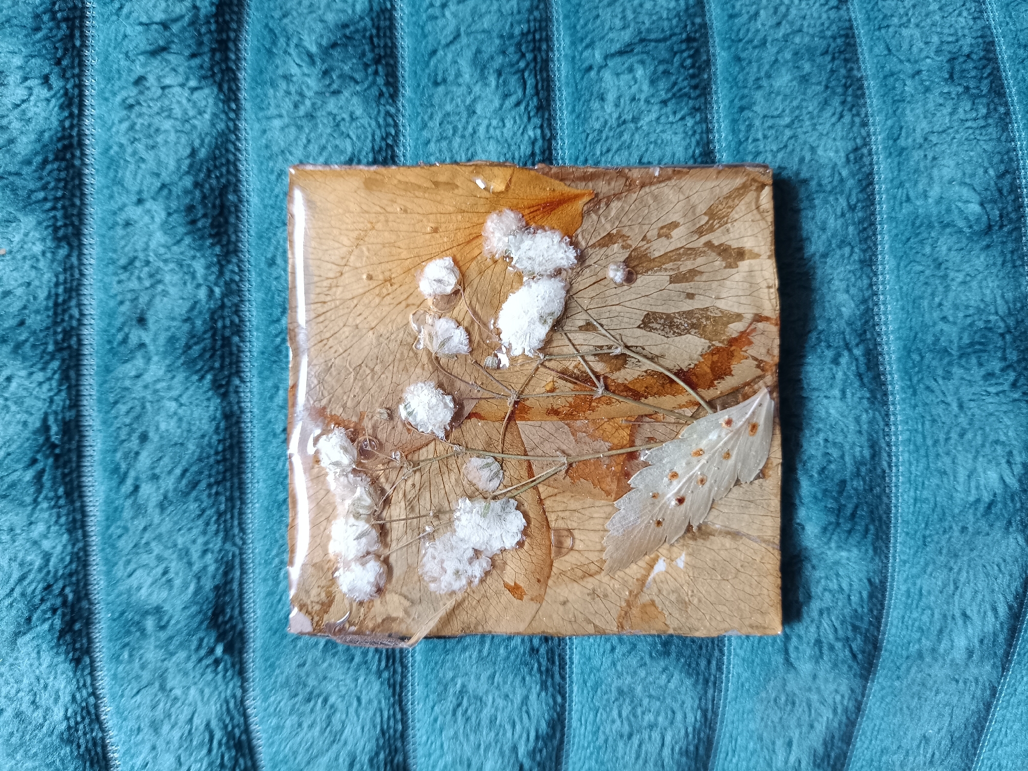 The image shows a square resin coaster with embedded dried leaves and white, fluffy decorations. The coaster is placed on a teal-colored, plush, textured surface, possibly a blanket or fabric backdrop. The leaves inside the coaster are various shades of brown and transparent. No people, actions, or interactions are visible in the image. There is no indication this image comes from Посад, Russia.