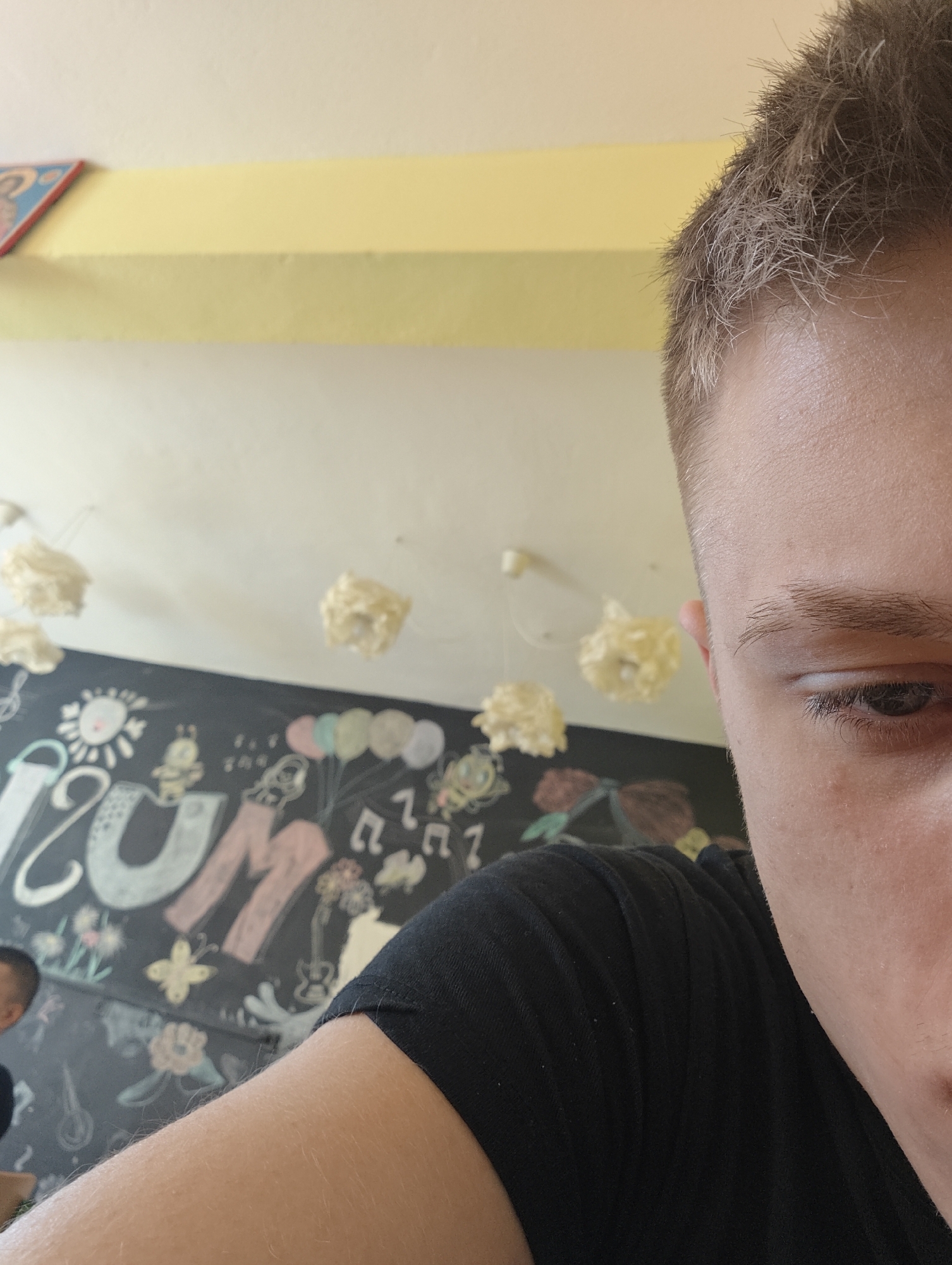 A close-up selfie taken indoors. The right side of a person's face is prominently displayed, showing part of their forehead, eye, and nose. They have short, light brown hair. They are wearing a black t-shirt. In the background, there is a black chalkboard with colorful drawings and writing. Above the chalkboard, the wall is white, with several white, flower-shaped light fixtures hanging from the ceiling. A yellow stripe is at the top of the wall. It is unknown if the image was taken in Kozloduy, Bulgaria.
