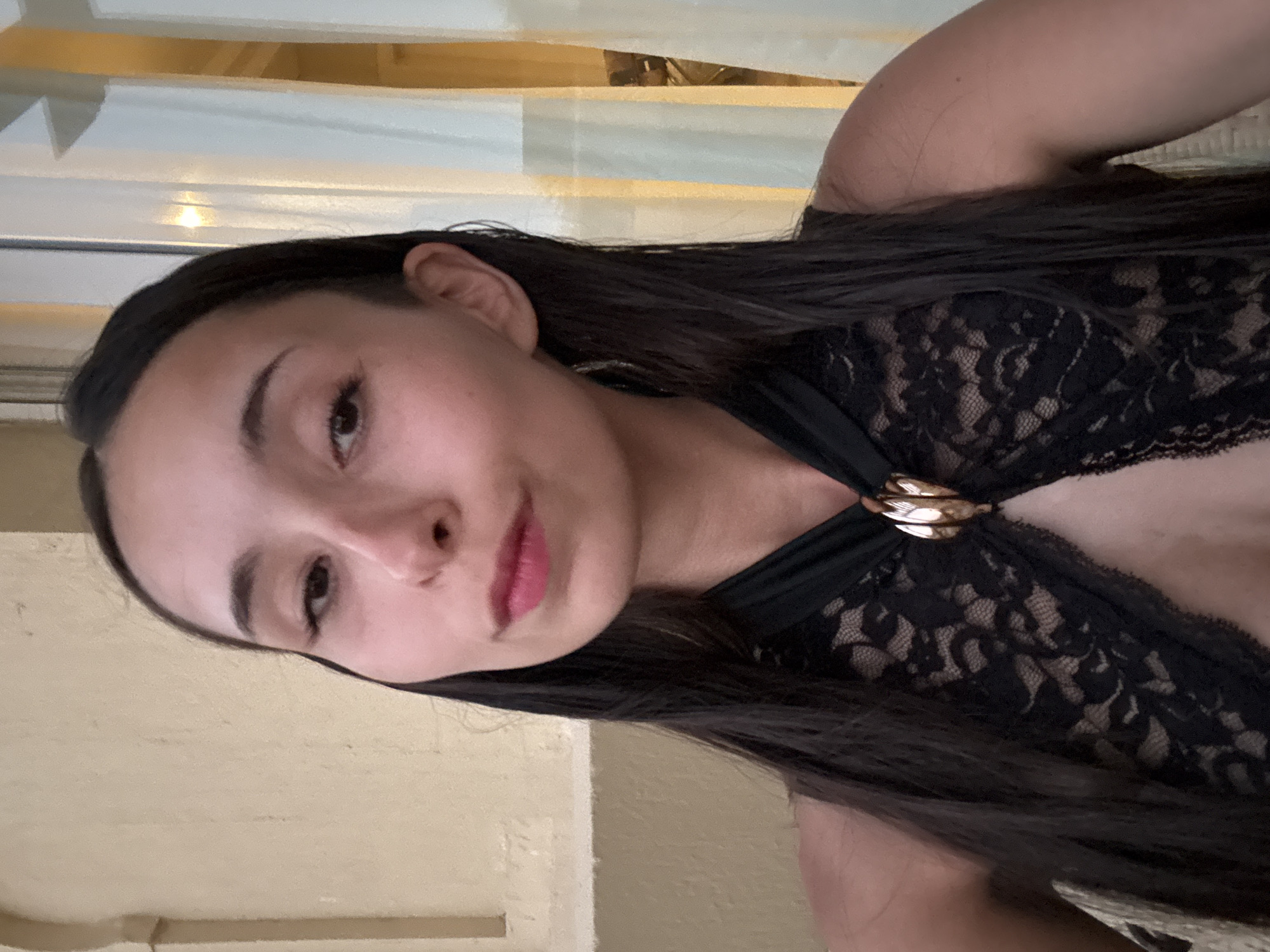 The image is a medium shot portrait of a young woman with long, dark, wet hair, looking directly at the camera. She is wearing a black lace top with a halter neckline that is adorned with a gold-colored brooch. Her makeup includes pink lipstick. The background is slightly blurred, showing a light-colored wall with architectural details and sheer curtains with a warm glow behind them, suggesting an indoor setting at dusk or night, possibly a hotel or restaurant. The overall impression is of a posed portrait taken during an evening outing. The location is specified as Unknown, Egypt.