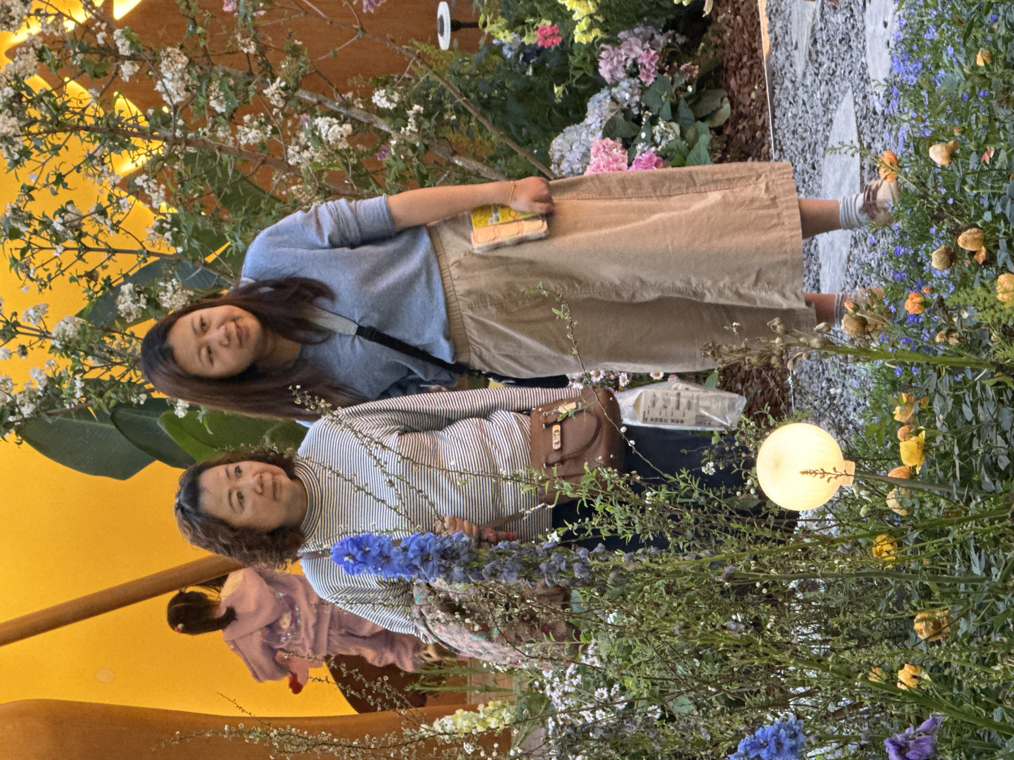 Two women are posing for a photo amidst a floral display. The woman on the left is wearing a striped shirt, while the woman on the right is wearing a blue sweater and tan pants. They are positioned among various flowers and plants, including blue delphiniums and white flowers. A lamppost with a spherical light fixture is also visible in the display. In the background, there is a yellow wall or structure. The photo was taken in the city of Liangjiang, China.