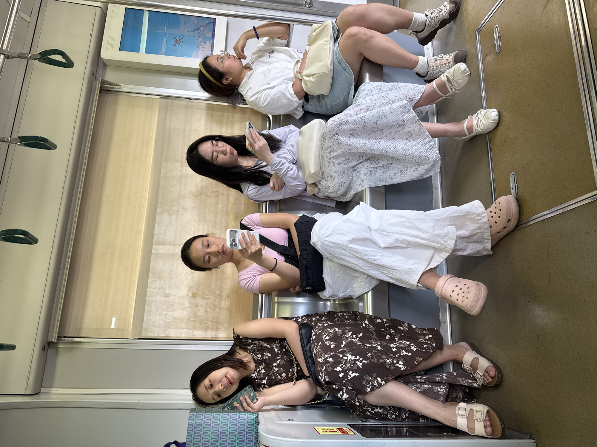 Four young women are seated on a public transport bench, likely a subway or train, in the Yuzhong District of China. The woman on the far left, with shoulder-length dark hair, is leaning back with her eyes closed and holding a green smartphone to her ear. She is wearing a brown floral-print dress and light-colored sandals. Next to her, a woman with dark hair tied back is looking down at her white smartphone. She is wearing a pink t-shirt and white loose-fitting pants, with a black bag slung over her shoulder. The third woman from the left has long dark hair and glasses, and she is also looking at her phone. She is wearing a light purple long-sleeved top and a white floral-print skirt, with a cream-colored bag on her lap. The woman on the far right has dark hair with a yellow headband and glasses. She is wearing a white collared shirt and a light blue denim skirt, and she is looking off to the side with her hand resting on her chin. A cream-colored bag is on her lap. The environment appears to be indoors with a window behind the women showing a diffused light, possibly a roller blind. The floor is a light brown color.