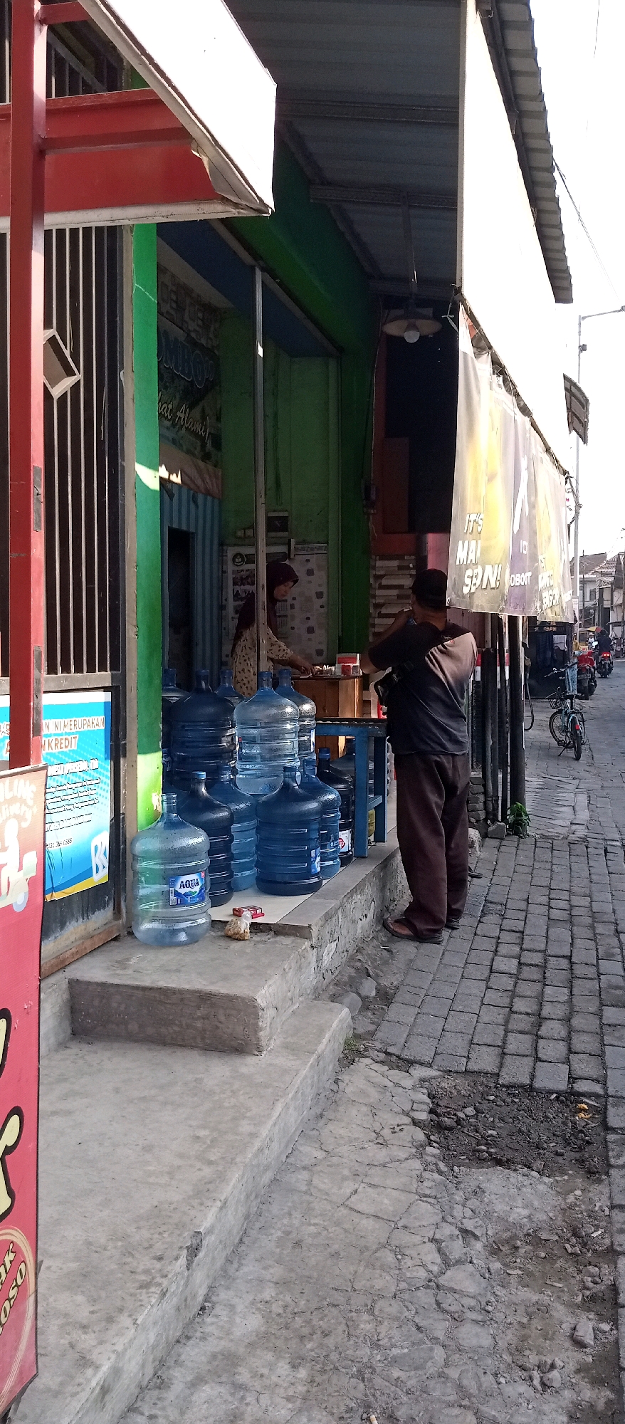 This image depicts a street-side stall in Wadungasri, Indonesia, likely a convenience store or a small food stall. The stall features a prominent display of large blue water jugs, some branded "AQUA," arranged on concrete steps and a blue metal table. In the background, a woman wearing a hijab and a patterned shirt is visible behind the counter, tending to the stall. She appears to be interacting with items on the counter, possibly preparing something for a customer or stocking. A man in a dark shirt and brown pants stands to the right of the stall, facing away from the camera, seemingly in the process of interacting with someone at the stall. He is wearing a dark cap and brown shoes. The stall itself is colorful, with green and red accents, and has a corrugated metal roof. Several banners or tarps hang from the front of the stall, some displaying text and graphics, one appearing to be a promotional banner with yellow and purple elements. The pavement in front of the stall is made of interlocking grey bricks, with some areas showing signs of wear and damage. A bicycle is parked nearby, and a few motorbikes are visible further down the street, suggesting this is an active area. The overall scene suggests a typical daytime street-level commercial activity in an Indonesian city.