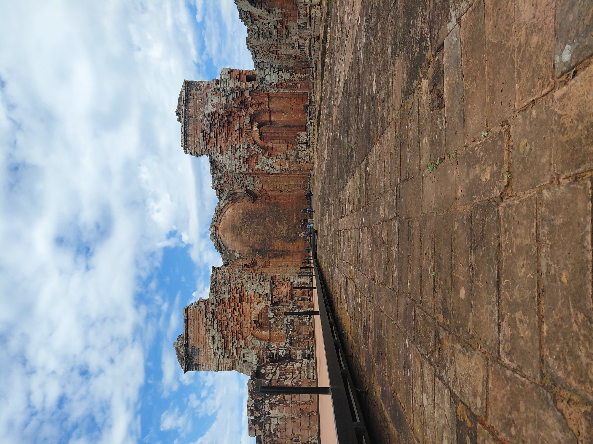 The image shows the ruins of a Jesuit mission in Trinidad, Paraguay. The ruins are made of red brick and are partially collapsed. There is a walkway in the foreground of the image, leading to the ruins. A few people are visible in the distance, walking around the ruins. The sky is blue with white clouds. The ground is paved with bricks.