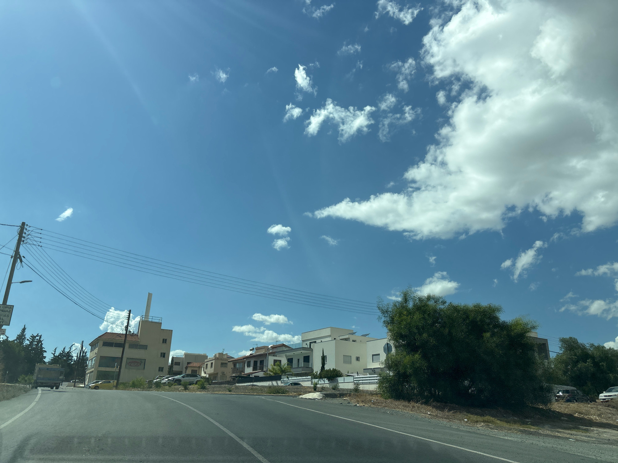 The image captures a street scene in Agios Athanasios Municipality, Cyprus, under a bright, partly cloudy sky. The perspective is from a moving vehicle, looking towards a row of buildings on the right side of the road. On the left, a utility pole with numerous wires runs parallel to the street, and a large truck is visible in the distance.
The buildings on the right range from a multi-story commercial building with a distinctive angled facade and a prominent chimney-like structure, to more modern, multi-level residential homes with flat roofs and white exteriors. There are several parked cars along the street and in front of the residences. A large, bushy green tree is prominently positioned in the foreground on the right, partially obscuring some of the buildings. The road itself is paved and appears to be a two-lane street with clear lane markings. The environment is dry and arid, with sparse vegetation along the roadside. The sun is casting strong shadows, indicating it is midday.