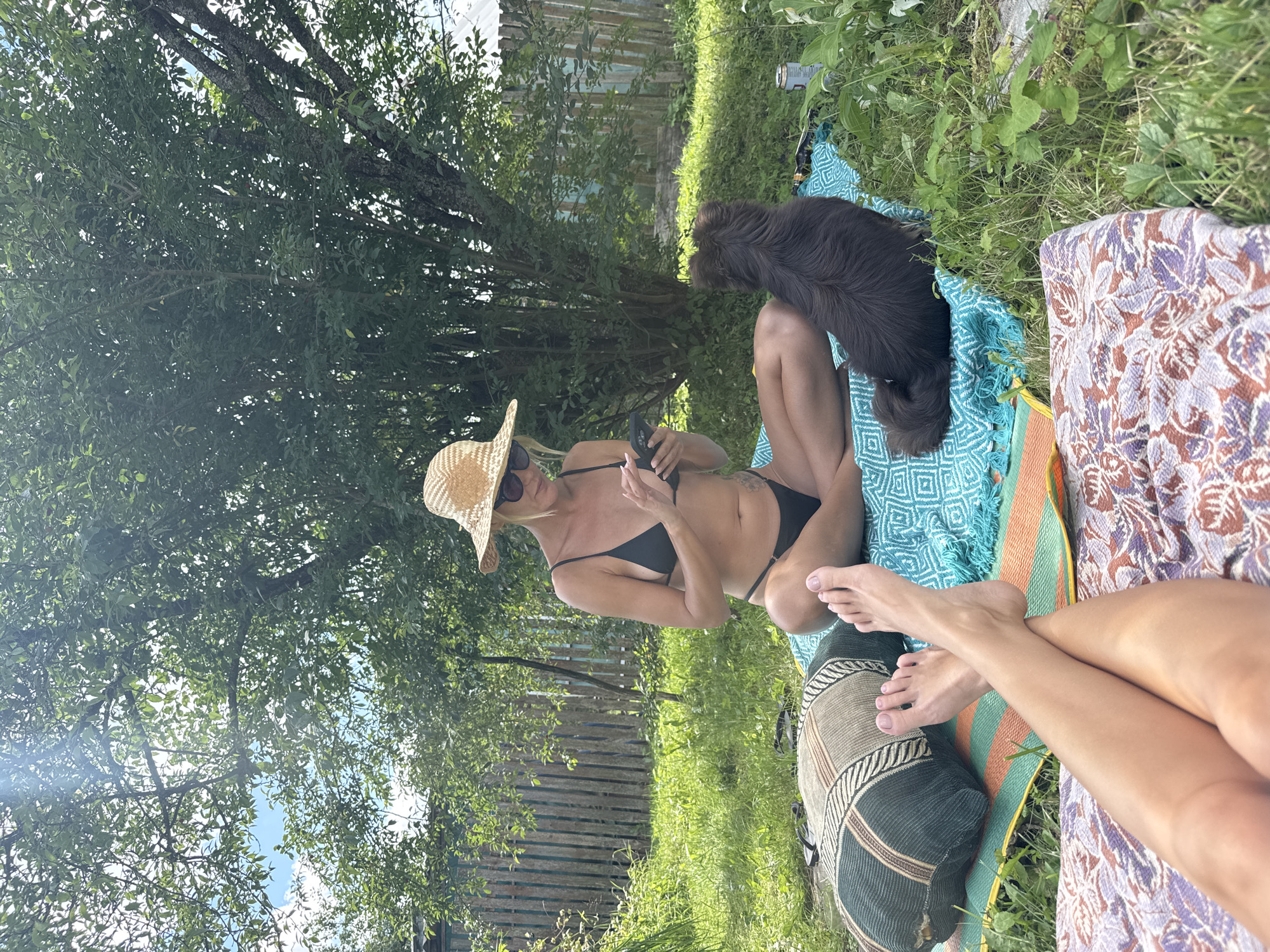In the SNT Krasnaya Presnya, Russia, a woman in a black bikini and a straw hat sits cross-legged on a turquoise blanket in a grassy backyard, looking at her phone. A small, fluffy brown dog sits beside her, facing away from the camera. In the foreground, the legs and feet of another person are visible, resting on patterned blankets. The scene is set under a leafy tree, with a wooden fence and rural buildings in the background. The lighting suggests it is a sunny day.
