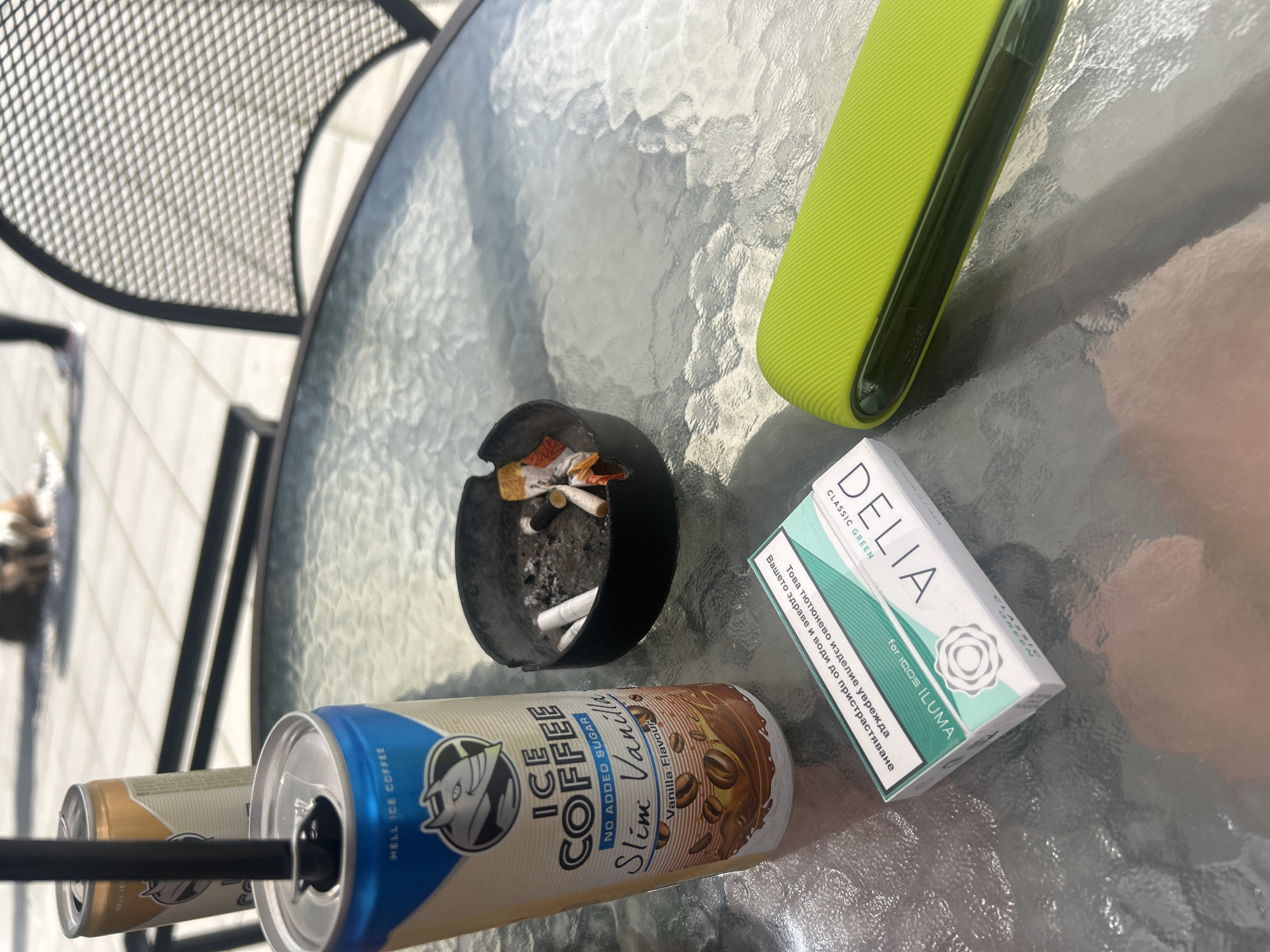 On a textured glass table top, an ashtray with cigarette butts is positioned near a pack of "Delia" Classic Green for IQOS ILUMA cigarettes. A green IQOS device rests nearby. Two cans of "Hell Ice Coffee," one blue and one gold, are visible, with a straw in the blue can. The scene appears to be outdoors, possibly in Plovdiv, Bulgaria, given the Cyrillic text on the cigarette pack. A metal chair is partially visible.